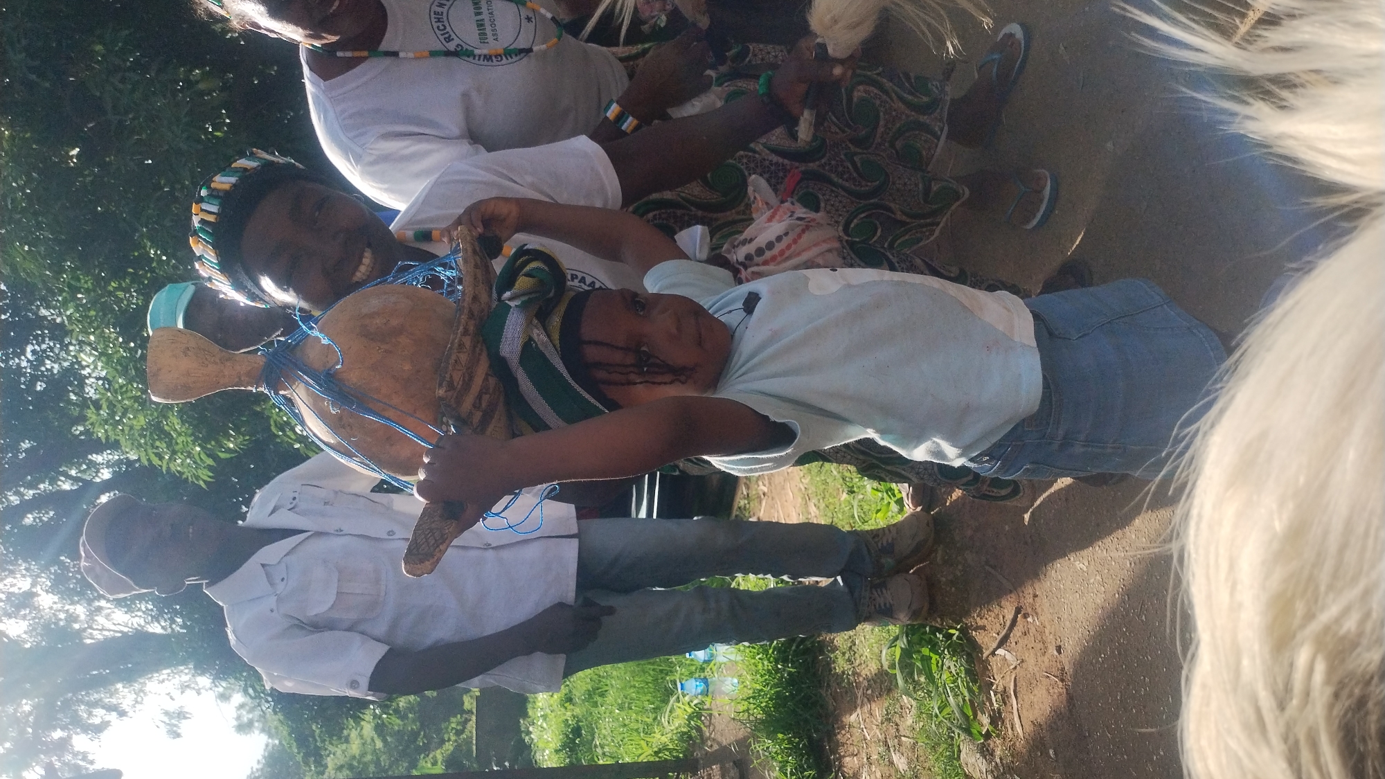 In Jos, Nigeria, a young child, positioned in the foreground and center, is hoisting a traditional gourd on their head, balancing it with their raised arms. The child wears a light blue t-shirt, a blue jean skirt, and a patterned headwrap with green and white stripes. Behind the child, a woman with a broad smile and a colorful beaded headband is visible, also wearing a white t-shirt. To the left, a man in a white collared shirt and light blue jeans stands, wearing a beige cap. The background reveals lush green trees and vegetation, with a dirt path underfoot, suggesting an outdoor, possibly rural or semi-urban, setting within Jos. The overall scene appears to capture a moment of cultural practice or celebration.