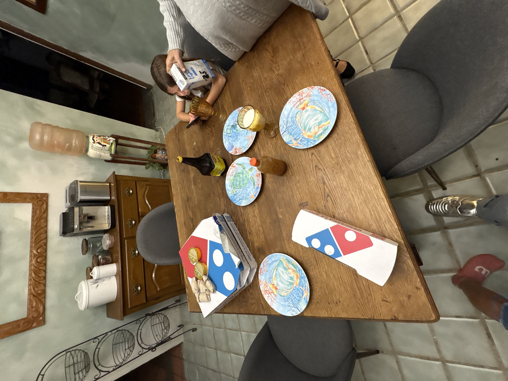 Here is a description of the image:
The image shows an angled overhead shot of a wooden dining table set with plates, food, and beverages, suggesting a casual meal setting. A child is present, seemingly pouring something from a carton into a glass. There is a person seated near the child, along with other individuals whose legs and feet are partially visible. The presence of a Domino's pizza box indicates that pizza is part of the meal. The environment seems to be indoors, likely a home dining area. The plates feature sea life themes.