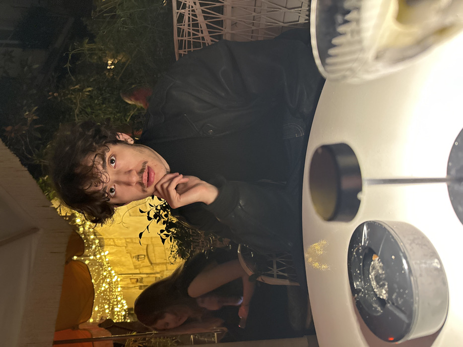 The image displays an evening scene on an outdoor terrace of a restaurant or bar in Paris, France. A young adult male, appearing to be in his twenties or early thirties, with dark, curly hair and a prominent mustache, is seated at a white table in the foreground. He is wearing a black turtleneck sweater and a black leather jacket, looking directly forward with a neutral expression. His right hand is positioned under his chin.
On the white table in front of him, a dark grey, oval-shaped ashtray containing cigarette butts and ash is visible. To its right, a small, dark-colored table lamp with a slender stem stands. A clear, patterned glass, possibly holding a drink, is partially visible on the far right.
In the blurred background to the left, another female individual with long, light brown hair is seated at an adjacent table, looking down at a glowing smartphone. Behind her, a light-colored fabric canopy is draped, illuminated by numerous warm-toned string lights. Beyond the canopy, an old stone building wall with a mounted sconce light is visible. To the right and behind the primary subject, dense green foliage and a white, geometric lattice structure are present. The overall scene is dimly lit, primarily by ambient and artificial light sources from the string lights and table lamp, indicating a nighttime environment.
