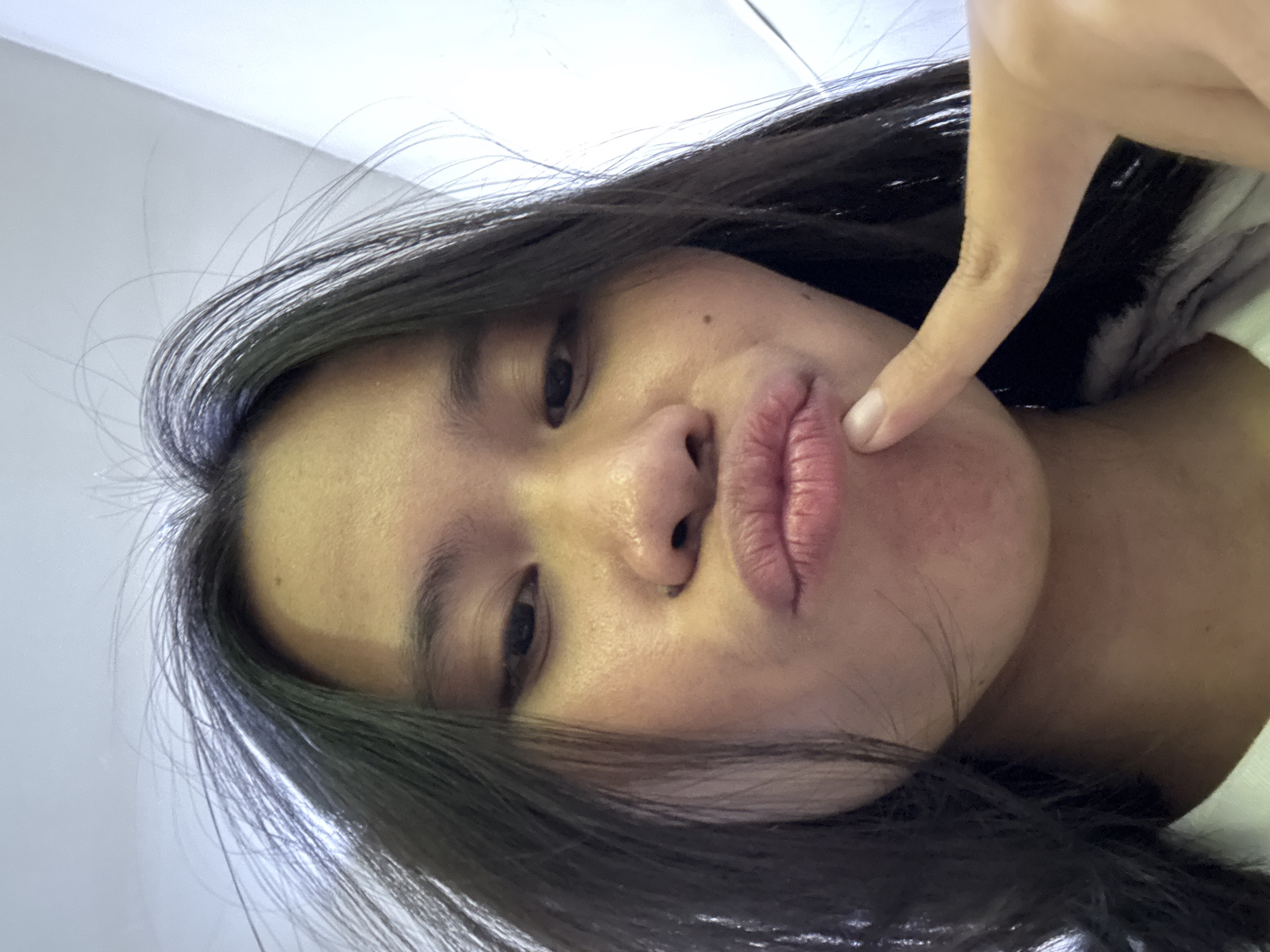 This is a close-up, waist-up selfie of a young woman with long, dark hair. She is looking at the camera with her lips pursed, and her right index finger is touching her chin. The background appears to be an indoor setting with white walls and a light fixture visible. The woman is wearing a light-colored shirt. The image does not contain any visual information that would indicate its location in East Jakarta, Indonesia.