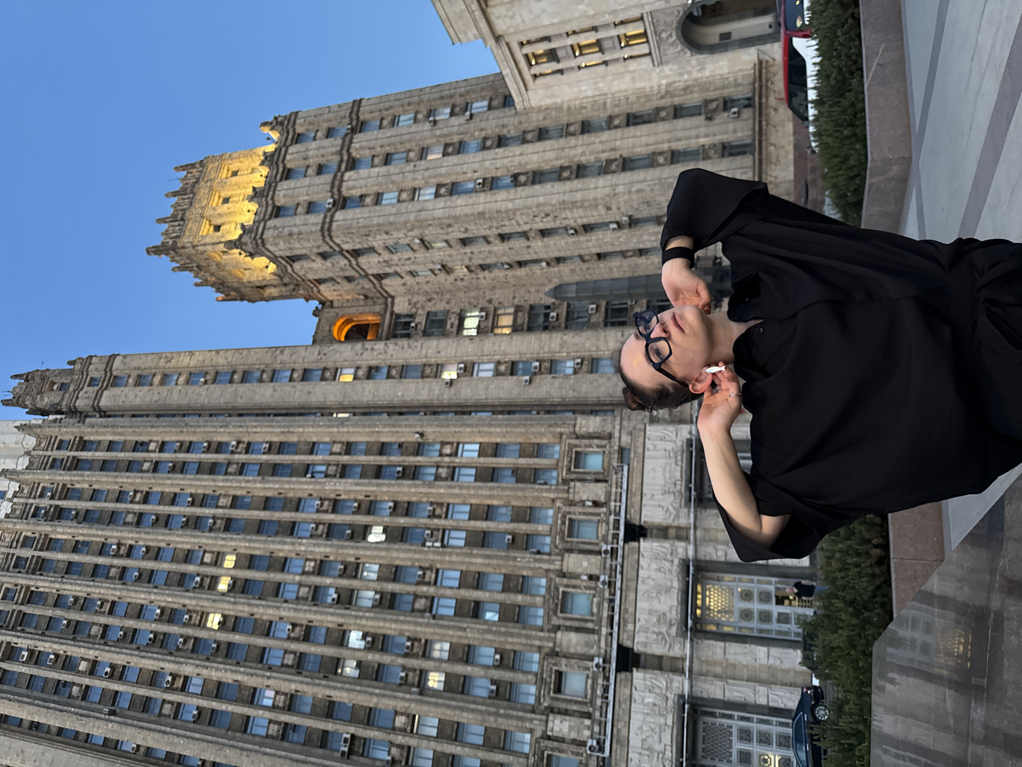 A woman in a black shirt and glasses is smoking a cigarette outside of the Seven Sisters high-rise building in Moscow, Russia. She is standing on a stone patio and the building is directly behind her.
