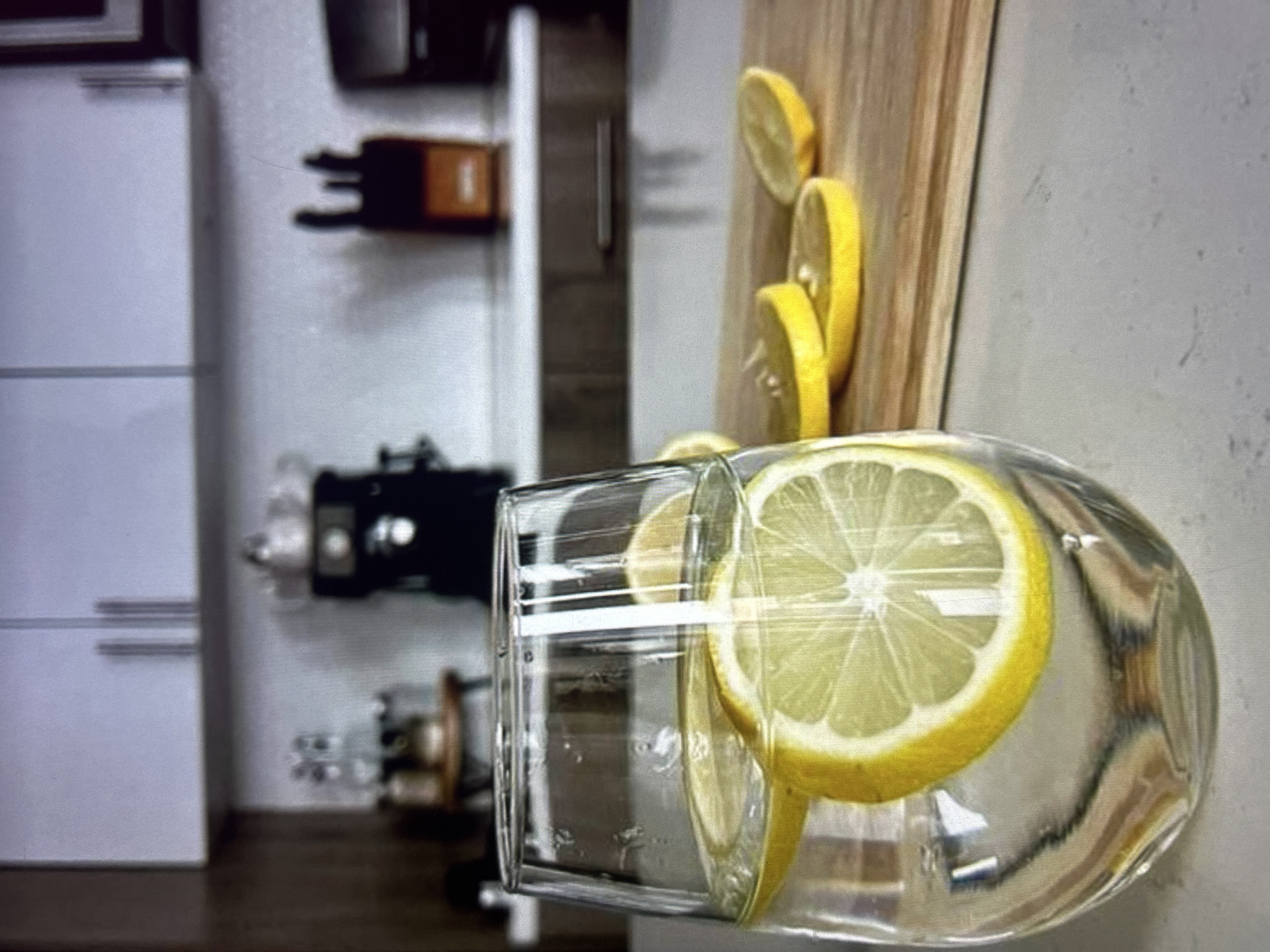 A clear glass of water with lemon slices sits on a countertop in a kitchen. Behind the glass, a wooden cutting board displays several more lemon slices. In the background, kitchen cabinets and appliances are visible, including a coffee maker and knife block. The scene appears to be set in a modern kitchen, possibly in Knezha, Bulgaria, given the context of the prompt. There are no people in the image.