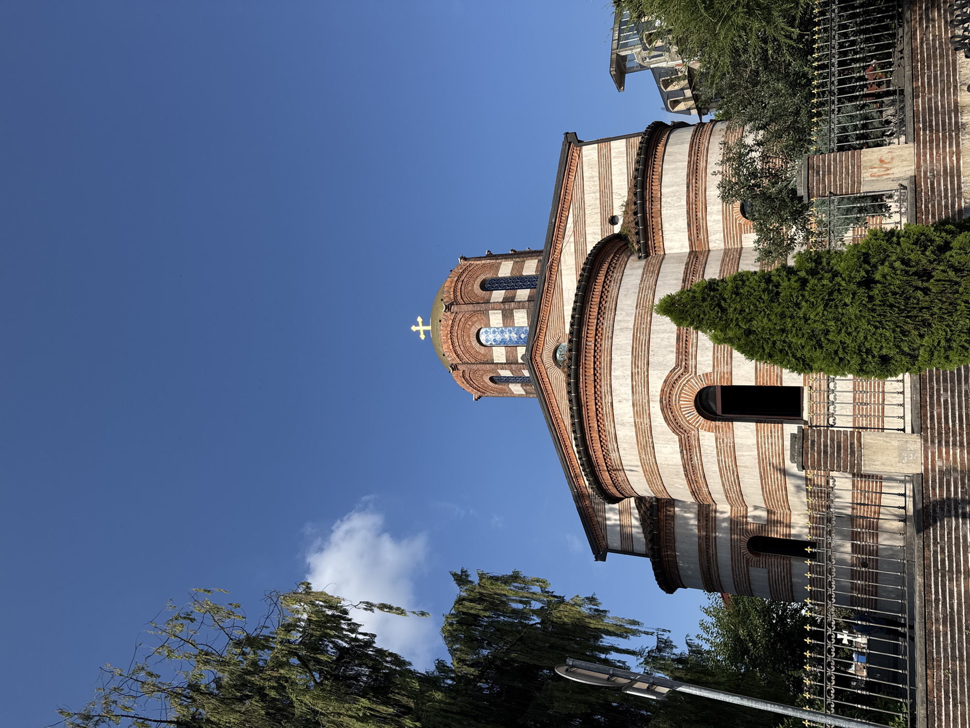 The image shows the Holy Trinity Church in Batumi, Georgia, under a clear blue sky. The church, a striking example of Byzantine architecture, features a golden dome topped with a cross. Its exterior is adorned with alternating bands of white stone and reddish-brown brick, creating a visually dynamic pattern. Several arched windows, some with stained glass, punctuate the facade.
To the left of the church, a large, leafy tree with drooping branches partially obscures the view, its foliage casting shadows. In the foreground, a decorative metal fence with gold accents runs along the perimeter, separating the church from the viewer. Behind the fence, a low brick wall and stone steps lead up to the church entrance, which is not fully visible. A young evergreen tree stands to the right of the entrance, adding a touch of greenery to the scene. In the background, beyond the church, modern buildings with glass facades suggest the urban setting of Batumi. The overall impression is one of a serene and historic religious site within a contemporary city.