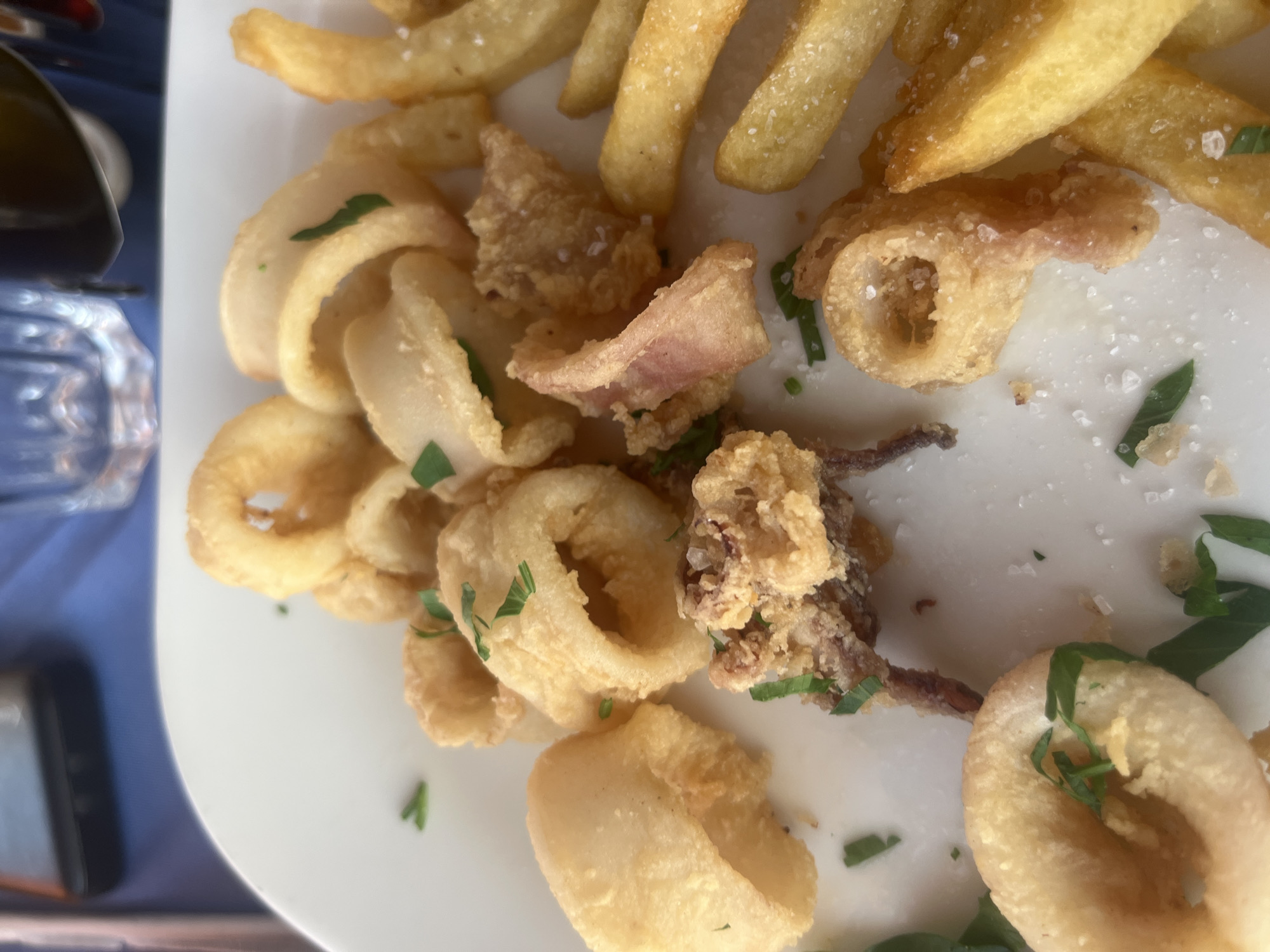 The image depicts a close-up view of a white plate filled with fried calamari rings and french fries. The calamari rings are golden brown and lightly seasoned with salt and chopped parsley. Some of the calamari pieces show tentacles. The french fries are also golden and appear to be seasoned with salt. The plate is set on a table with a blue tablecloth, and a glass of water and a dark object, possibly a phone, are visible in the blurred background. The setting appears to be a restaurant in Polis Chrysochous, Cyprus, suggesting a casual dining experience.