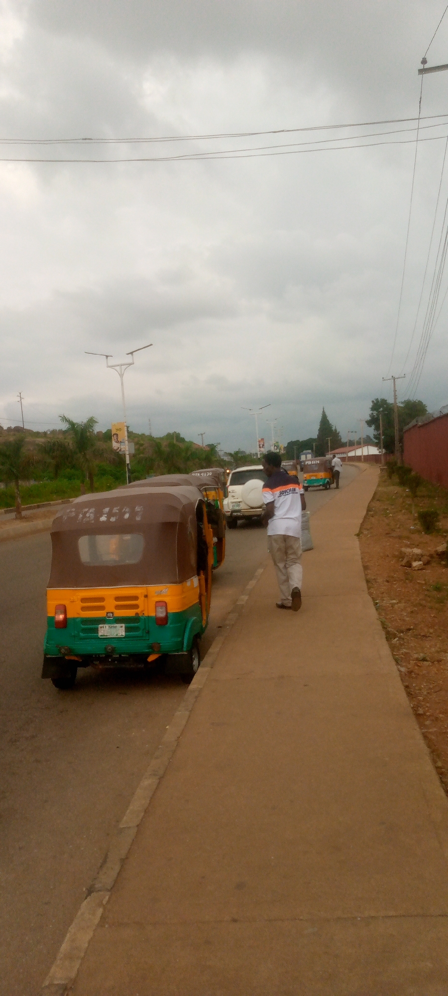 A cloudy day scene depicts a street with several three-wheeled vehicles, commonly known as auto-rickshaws, and a white SUV parked or driving on the road. A man wearing a white t-shirt with orange and black trim, and light-colored pants walks away from the camera on a paved sidewalk. The auto-rickshaws are colorful, with one in the foreground primarily green and yellow with a brown canopy, displaying a license plate that reads "TAA 4507". The sidewalk is a light brown color, bordered by a darker strip on the road's edge. In the background, there are trees, streetlights, and a red brick wall. The sky is overcast with a muted, grey hue.
