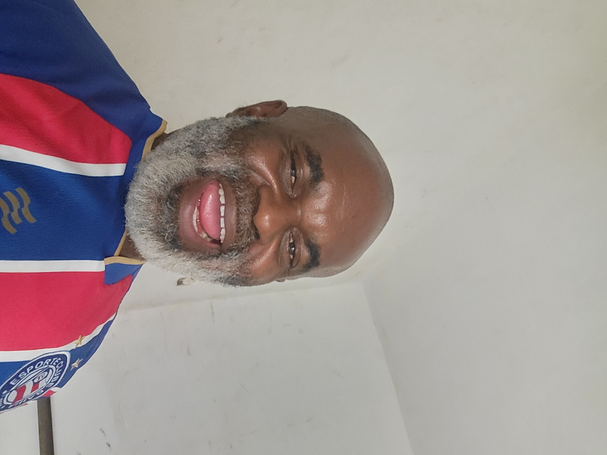 A portrait of a smiling man with a shaved head and a gray beard, wearing a blue, red, and white striped shirt with a logo on the left side. The background appears to be a plain white wall. The image is rotated. There is no indication in the image of the location in the city of Maiburiji, Nigeria.