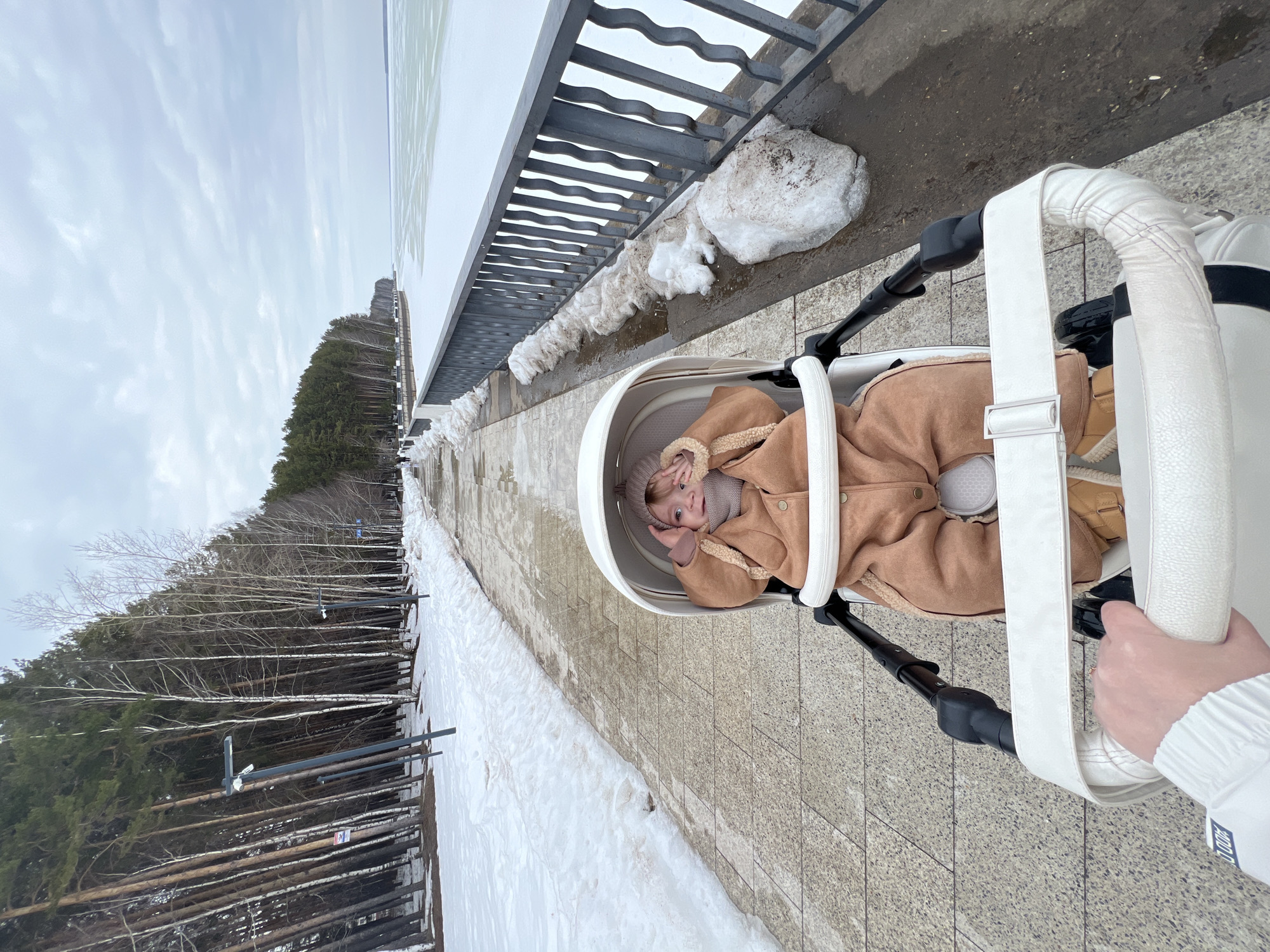 A photo taken in Chaikovsky, Russia, shows a baby lying in a white stroller. The baby wears a brown coat with a fur-trimmed hood and is looking up at the camera. The stroller is being pushed along a paved path next to a metal railing. Beyond the railing is a snow-covered area with trees. The sky is overcast. The baby's hand is visible holding the stroller's handle. Snow is on the ground next to the path. The baby is facing away from the railing.