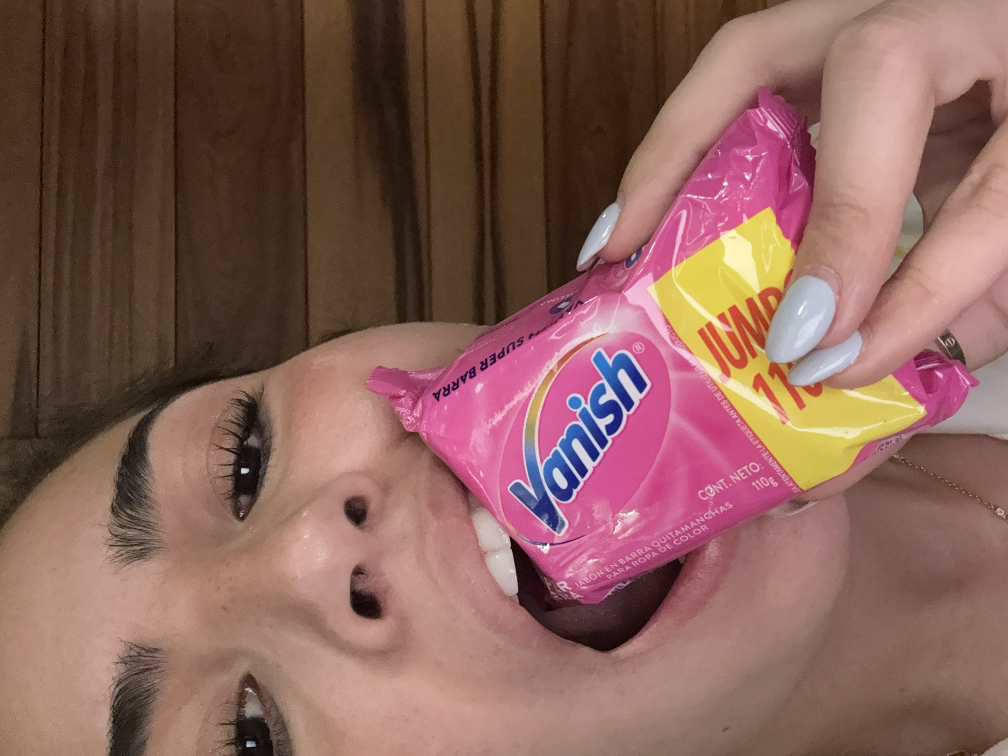 A woman lies on her side, facing the camera, with a pink and yellow packet of "Vanish" laundry stain remover partially inserted into her open mouth. She holds the packet with her right hand, her fingers adorned with light blue nail polish. The product's packaging includes the brand name "Vanish" in blue lettering. The text "SUPER BARRA" and "JUMBO 110g" are visible on the packaging. The background appears to be a wooden surface. The location is San Bernardino Tlaxcalancingo, Mexico.