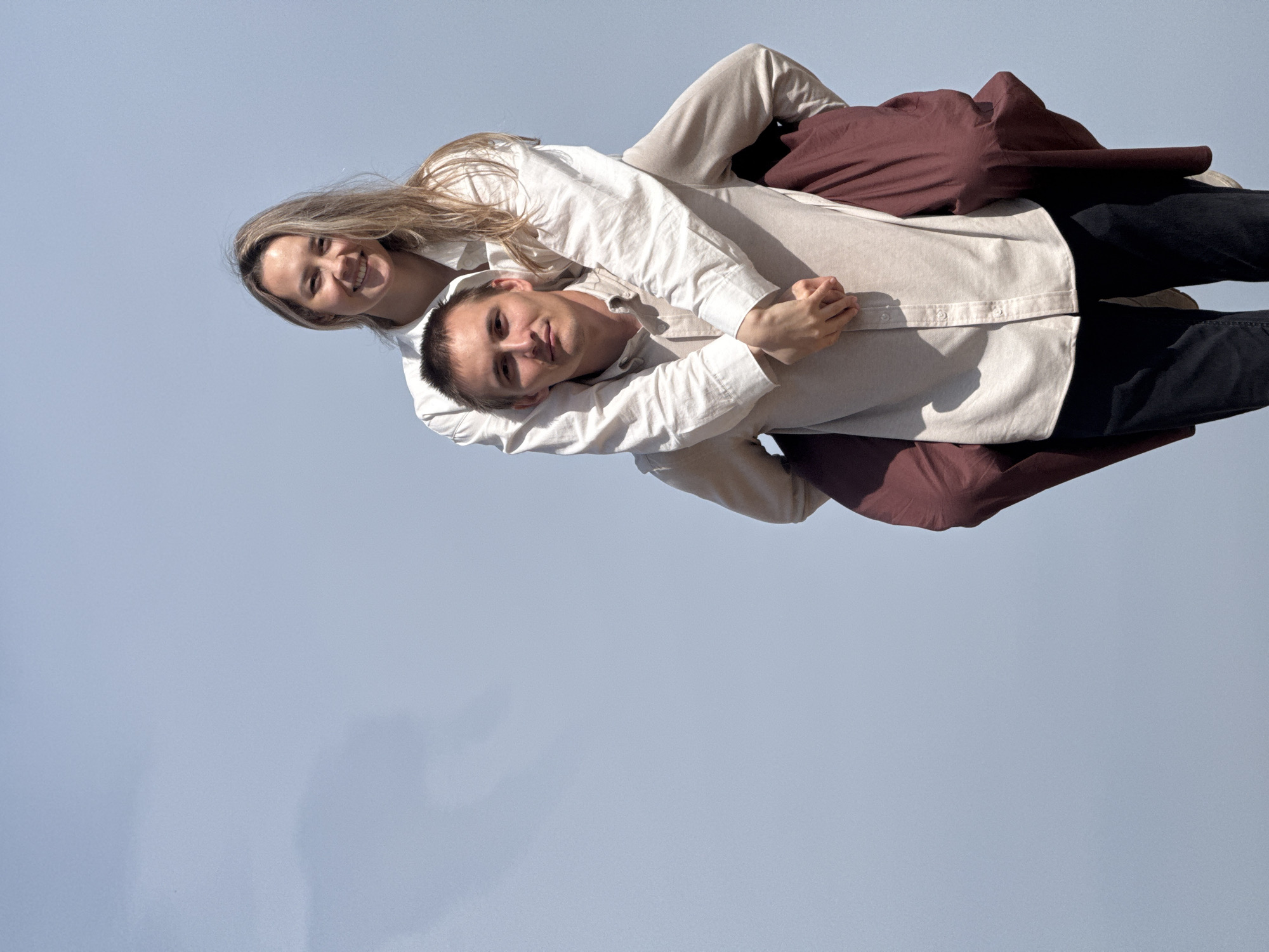 A couple poses against a light blue sky, likely outdoors in daylight. The woman, with blonde hair, is riding piggyback on the man, both wearing light-colored button-down shirts and smiling. The man is wearing black pants, and a reddish-brown garment drapes over him. The location is unidentified.