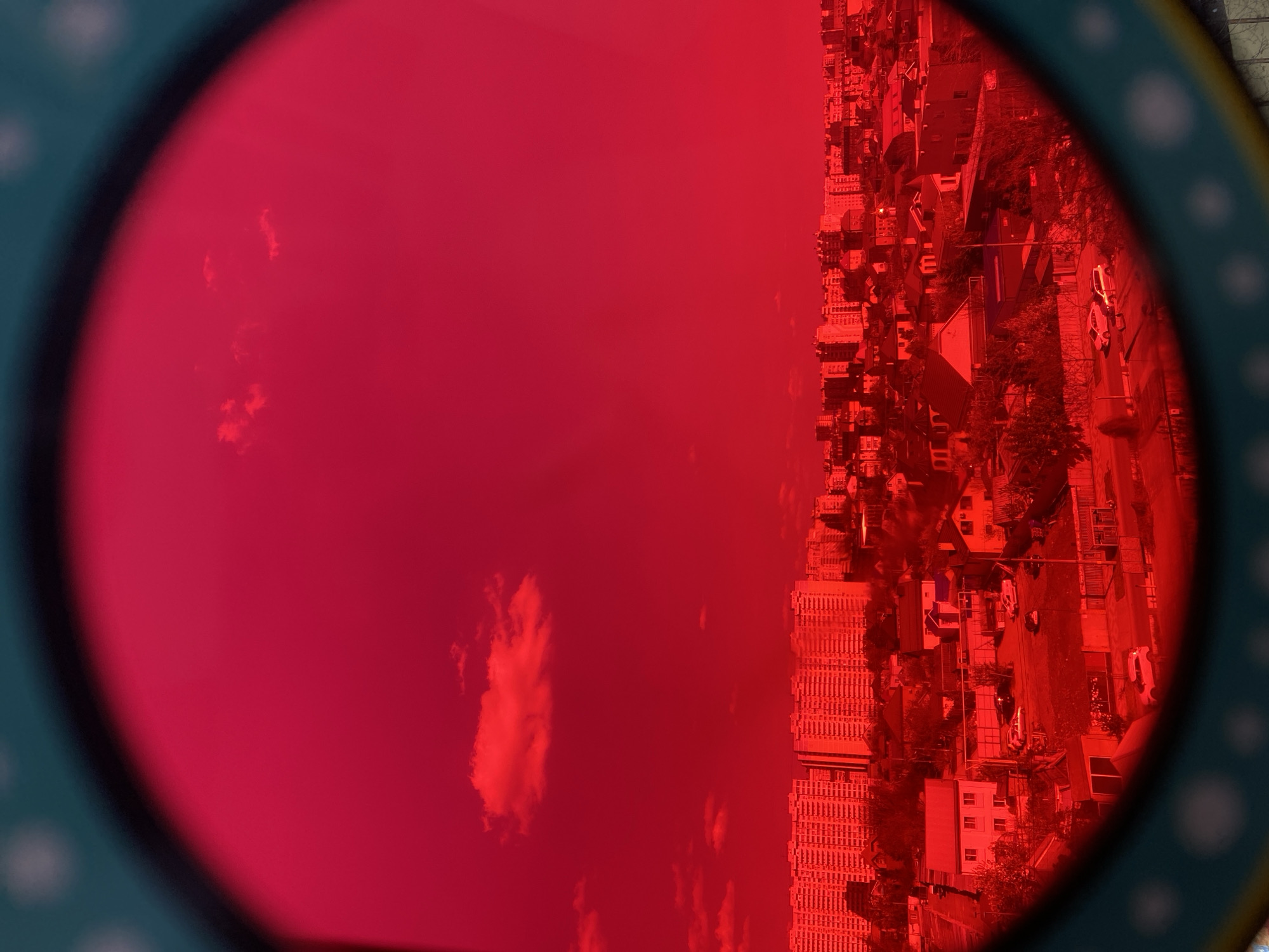The image displays an outdoor scene viewed entirely through a circular, red-tinted filter or lens. The frame of this filter is visible along the left and bottom-right edges, appearing dark teal or dark green with white polka dots.
Within the vibrant red circle, a city or town landscape is captured from a high vantage point. The overall image is presented in portrait orientation, but the perspective within the red circle suggests the scene itself is rotated approximately 90 degrees clockwise.
On the right side of the red-filtered view, a dense collection of buildings dominates the landscape. These include larger multi-story structures, likely commercial or residential buildings, and smaller, individual houses. Roads, some containing indistinct shapes that appear to be vehicles, weave between these structures. Patches of trees are also discernible, contributing to the urban or suburban setting. All elements of the cityscape are monochromatic, rendered in deep reds and darker crimson tones.
The left side of the red circle primarily shows the sky, featuring several clouds. These clouds appear as lighter, irregularly shaped red masses against the slightly darker red expanse of the sky. The extreme red tint obscures natural colors, making it impossible to determine the exact time of day or specific details like building materials. There is no visible text or identifiable landmarks within the scene.