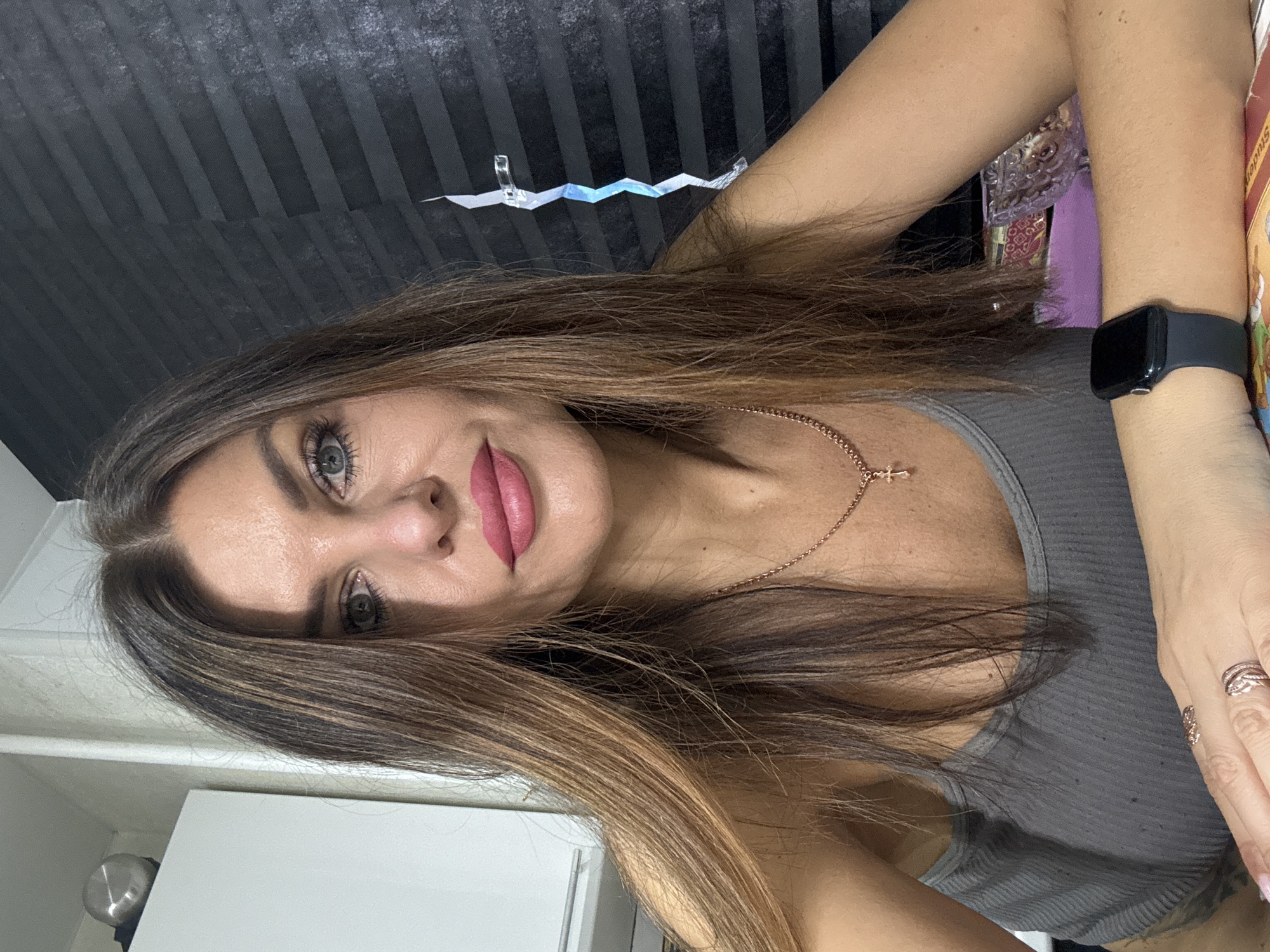 A woman with long brown hair and grey eyes is posing for a selfie. She is wearing a grey tank top and a gold necklace with a cross pendant. She has a black smartwatch on her left wrist and rings on her fingers. The background is a dark gray wall with horizontal lines and a white cabinet. There is also a small decorative item hanging above her head. The location is likely indoors. The specific city of Chaikovsky, Russia, is not discernible from the image.