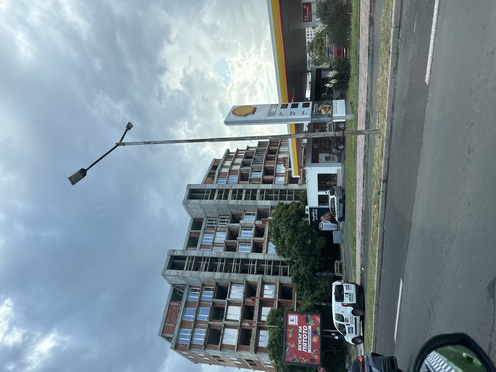 The image captures a cloudy day scene in Burgas, Bulgaria, with a focus on a partially constructed multi-story building on the left and a Shell gas station on the right. The building is in a state of construction, with exposed brickwork and concrete structures. Windows are installed in some parts, while others remain open. In front of the building, a billboard advertising "Kaufland" with images of fruits is visible.
Adjacent to the building is a Shell gas station, featuring its signature yellow and red branding. A prominent Shell sign stands tall. The gas station appears to be operational, with a covered canopy over the pumps and cars visible.
In the foreground, a street with traffic is depicted, suggesting the location is along a main road. A white van with signage is parked on the side of the road, and other vehicles are also present. The environment appears to be a mix of urban development, with the construction site and commercial establishments like the gas station. Lush green trees are situated between the building and the road. The overall atmosphere is somewhat overcast due to the cloudy sky.
