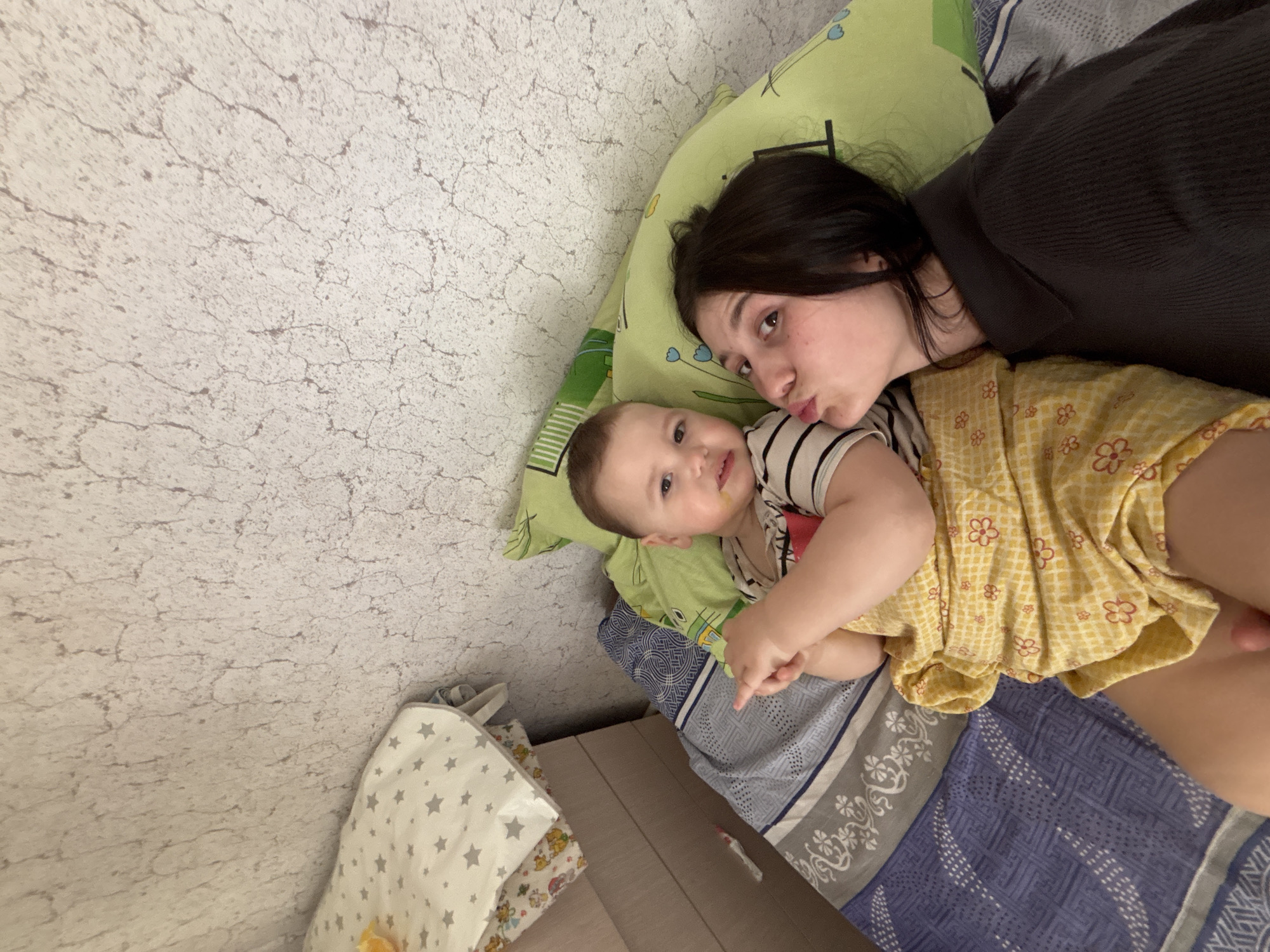 The image depicts a woman and a baby lying on a bed. The woman has dark hair and is wearing a dark shirt. She is looking at the camera with pursed lips. The baby is wearing a striped shirt and a yellow dress with a floral pattern. The baby is looking at the camera with a slight smile. The bed has a green pillow and a blue and gray patterned sheet. The background is a light-colored wall with a textured surface. There are also bags on the floor near the bed. The location is most likely Фоки, Russia, based on the prompt.
