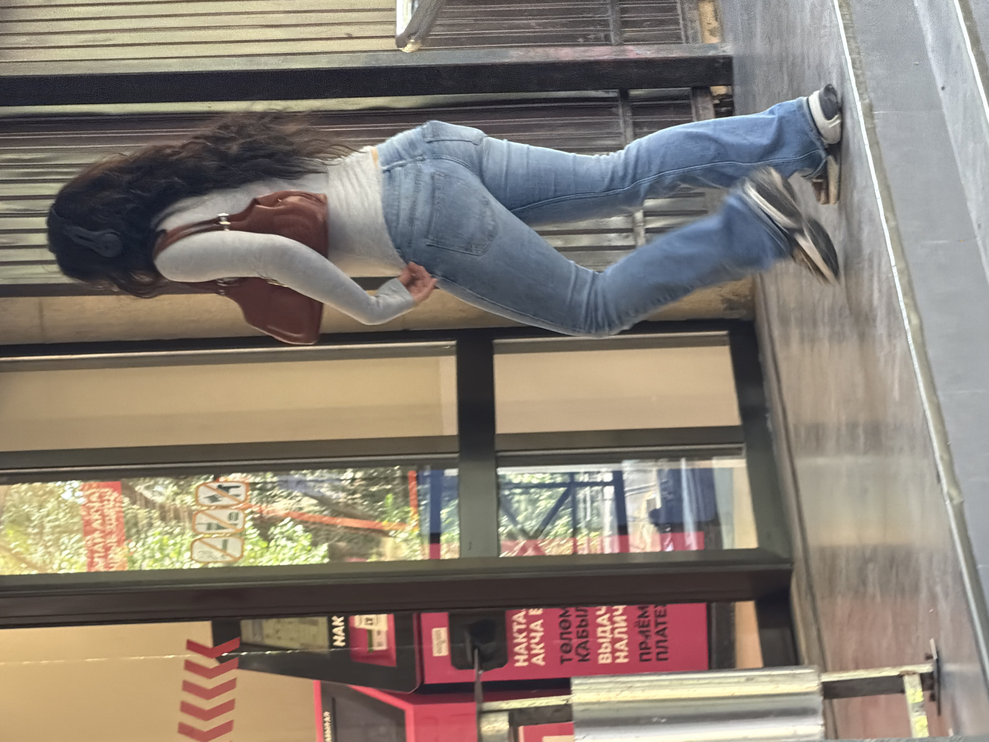 A medium shot captures a young woman from the back as she walks away from the camera, exiting a building through a glass door. She has long, dark, wavy hair and is wearing a light grey long-sleeved shirt, blue flared jeans, and white and brown athletic shoes. A brown purse hangs over her right shoulder. The background reveals a glimpse of trees through the glass, with an ATM machine visible to the left. Signs with red text and Cyrillic lettering are posted on the ATM. The woman's feet are blurred due to motion as she steps down. The setting appears to be an outdoor urban environment.