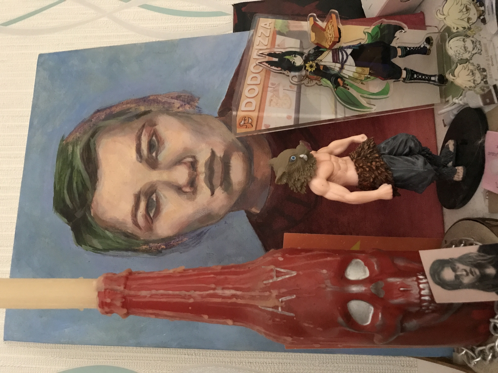 The image shows a close-up of a collection of objects on a surface. There is a painting of a person with green hair and light skin with blue paint in the background, a clear acrylic sign with the word "DODODIZZA" on it, an anime figure, a figurine of a man with a boar head, a skull-shaped candle with a portrait attached, and other miscellaneous objects. The location is unspecified.