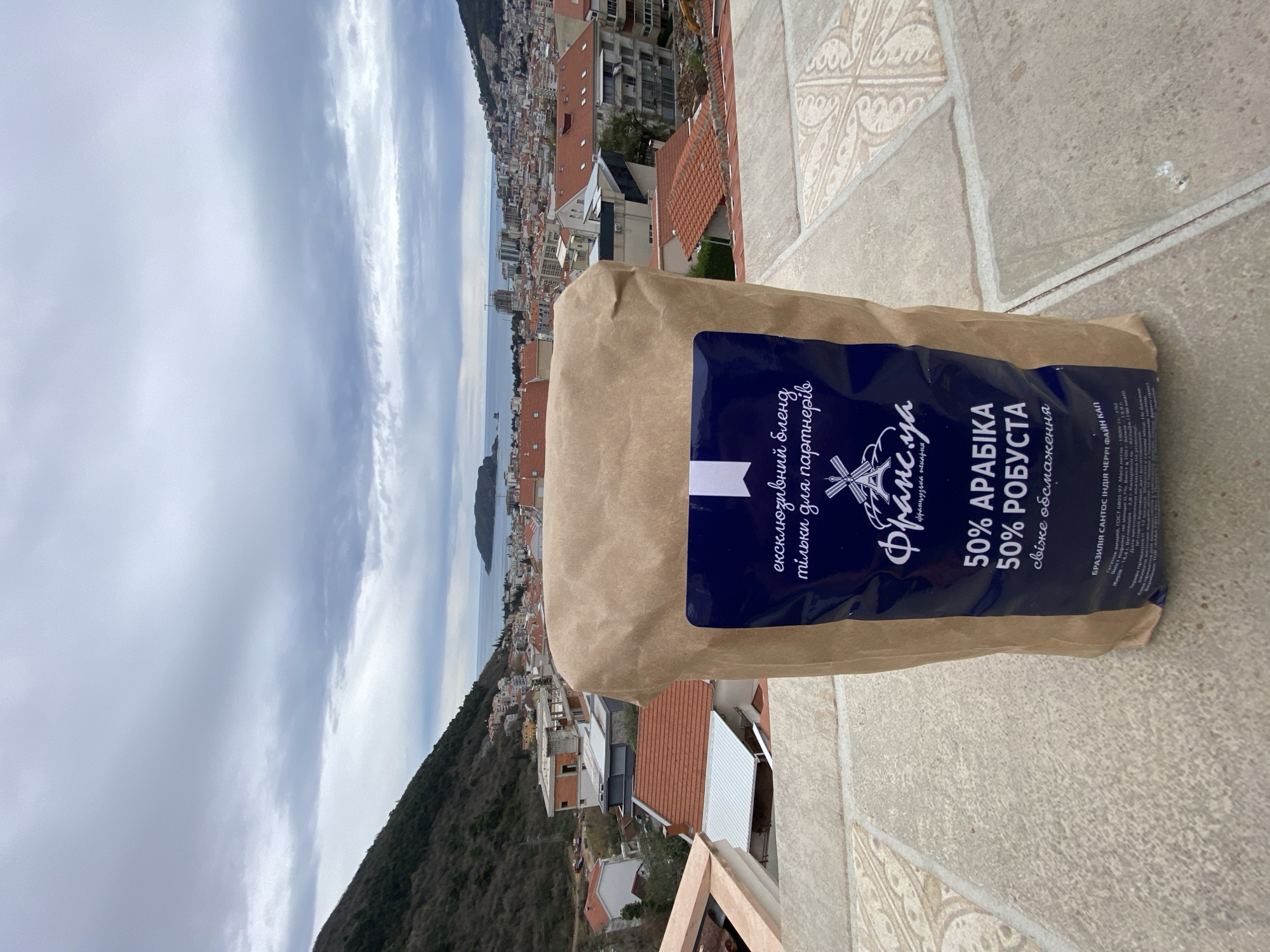 The image shows a brown paper bag of "Frantsiya" coffee blend (50% Arabica, 50% Robusta) sitting on a tiled surface. The packaging features a blue label with text in Ukrainian. In the background, there is a cityscape with buildings, red tile roofs, and trees, sloping down towards the sea. The sky is overcast with a view of the city of Budva, Montenegro and the mountains behind it. The scene is shot from an elevated perspective.