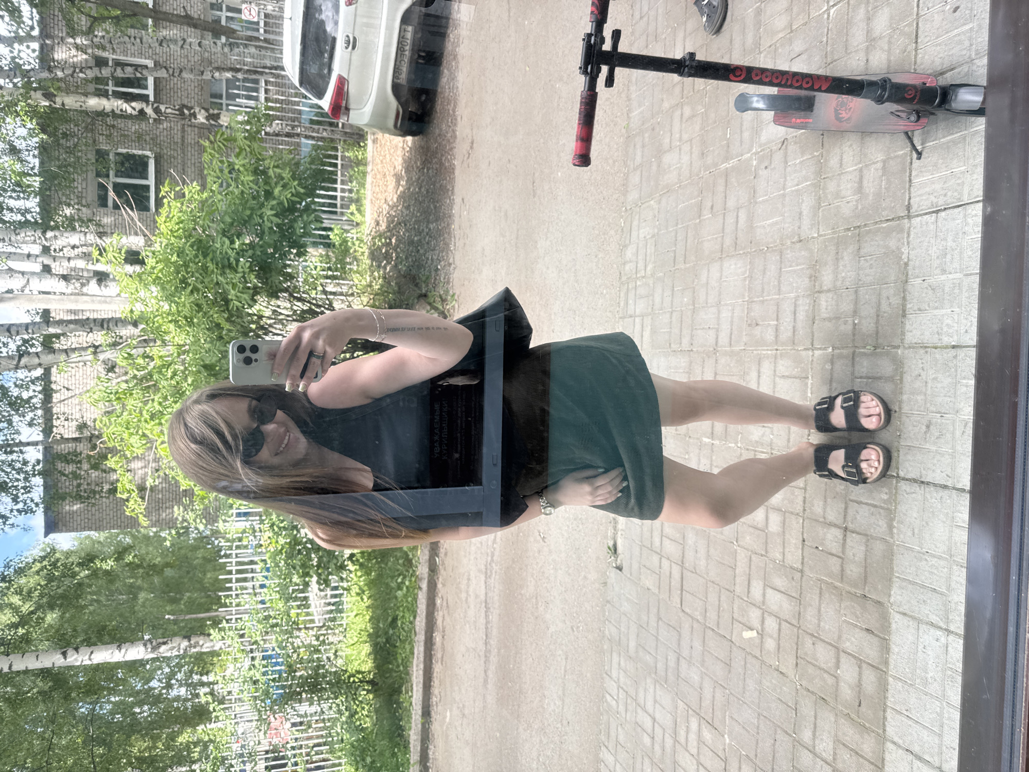A full-length mirror selfie of a woman in her late 20s to early 30s. She is wearing a black sleeveless top, a dark green skirt, and black sandals. She is holding her phone in her right hand, taking the photo. She has long, brown hair and is wearing sunglasses. She is smiling. In the background, there is a building, trees, a fence, a car, and a kick scooter. The scene appears to be in a residential area in Chaikovsky, Russia, judging by the architecture and vegetation.
