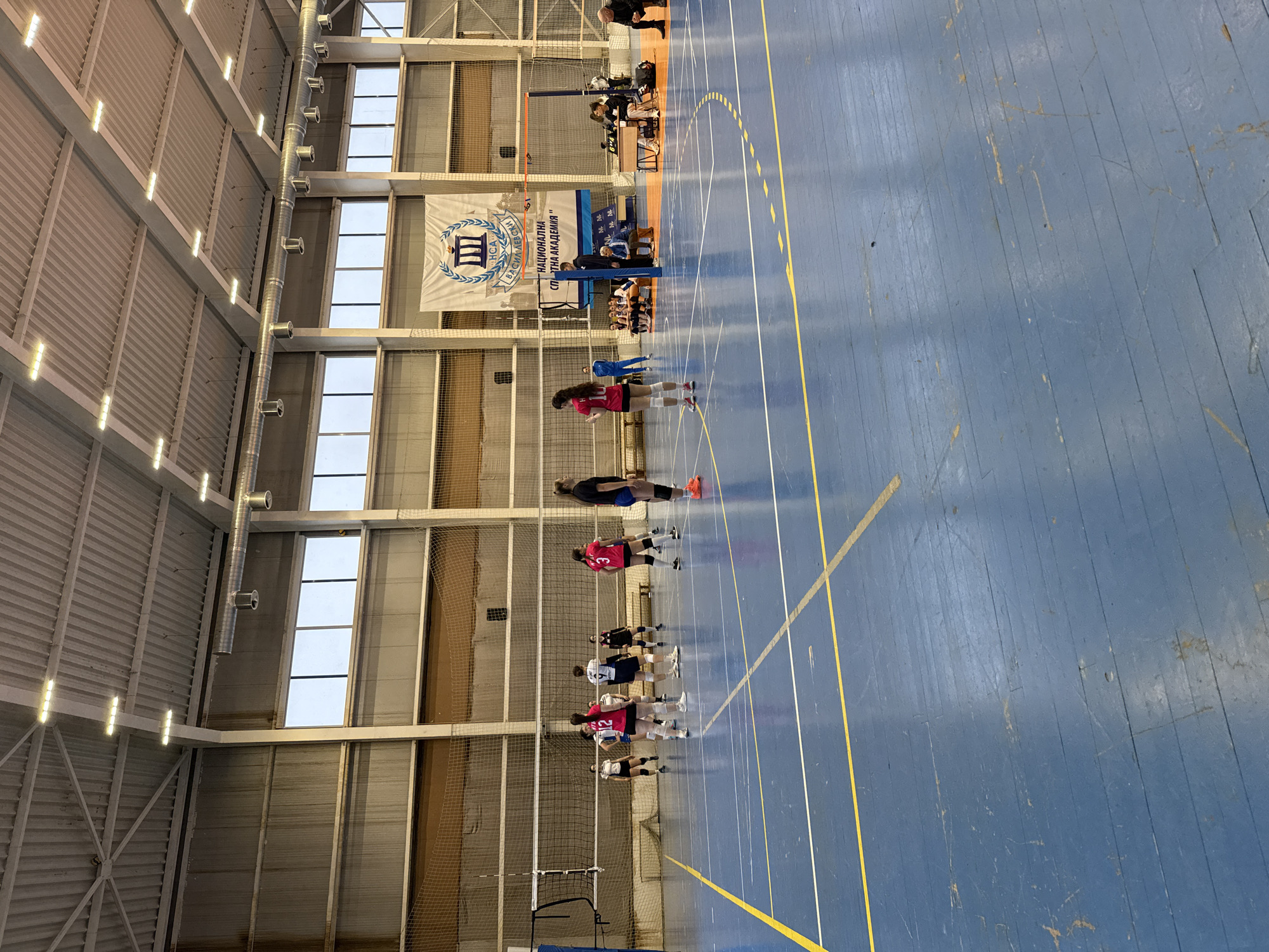 The image captures an active indoor volleyball court within a gymnasium at the National Sports Academy "Vasil Levski" in Sofia, Bulgaria. The court floor is composed of blue-painted wooden planks, marked with yellow and white lines defining volleyball boundaries. A white net with blue support poles divides the court. Approximately ten to twelve female athletes are present; several wear pink/red jerseys with numbers (e.g., 10, 12, 3), while others wear dark blue/black jerseys (e.g., 9). An orange training cone rests on the court near the center. To the right of the net, a scorekeeping table features an electronic display showing "8" and "4," attended by an individual in a dark shirt and light pants. Multiple individuals, including athletes and presumed coaches or officials, are seated on benches along the right sideline. The gymnasium ceiling is high, constructed of corrugated metal panels, equipped with multiple linear LED light fixtures and exposed cylindrical ventilation ducts. Large rectangular windows are visible along the upper wall. A prominent banner hanging on the far wall identifies the venue as "НСА ВАСИЛ ЛЕВСКИ" (NSA Vasil Levski) and "НАЦИОНАЛНА СПОРТНА АКАДЕМИЯ" (National Sports Academy). The walls above court level are covered with protective netting.