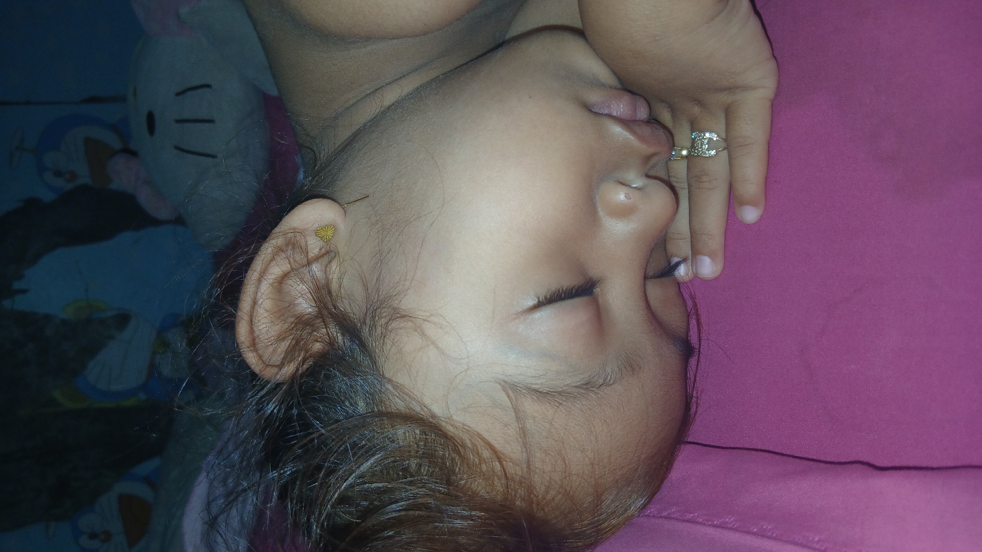 In Keudejungkagajah, Indonesia, a close-up, overhead shot captures a young child asleep on a pink surface, likely a bed or blanket. The child's face is turned to the right, with eyes closed and mouth slightly ajar. A golden heart-shaped earring is visible in the child's left ear. The child's right hand rests on their cheek, adorned with a ring featuring multiple small stones. The background is dimly lit and blurred, but suggests the presence of stuffed toys, including one resembling Doraemon.