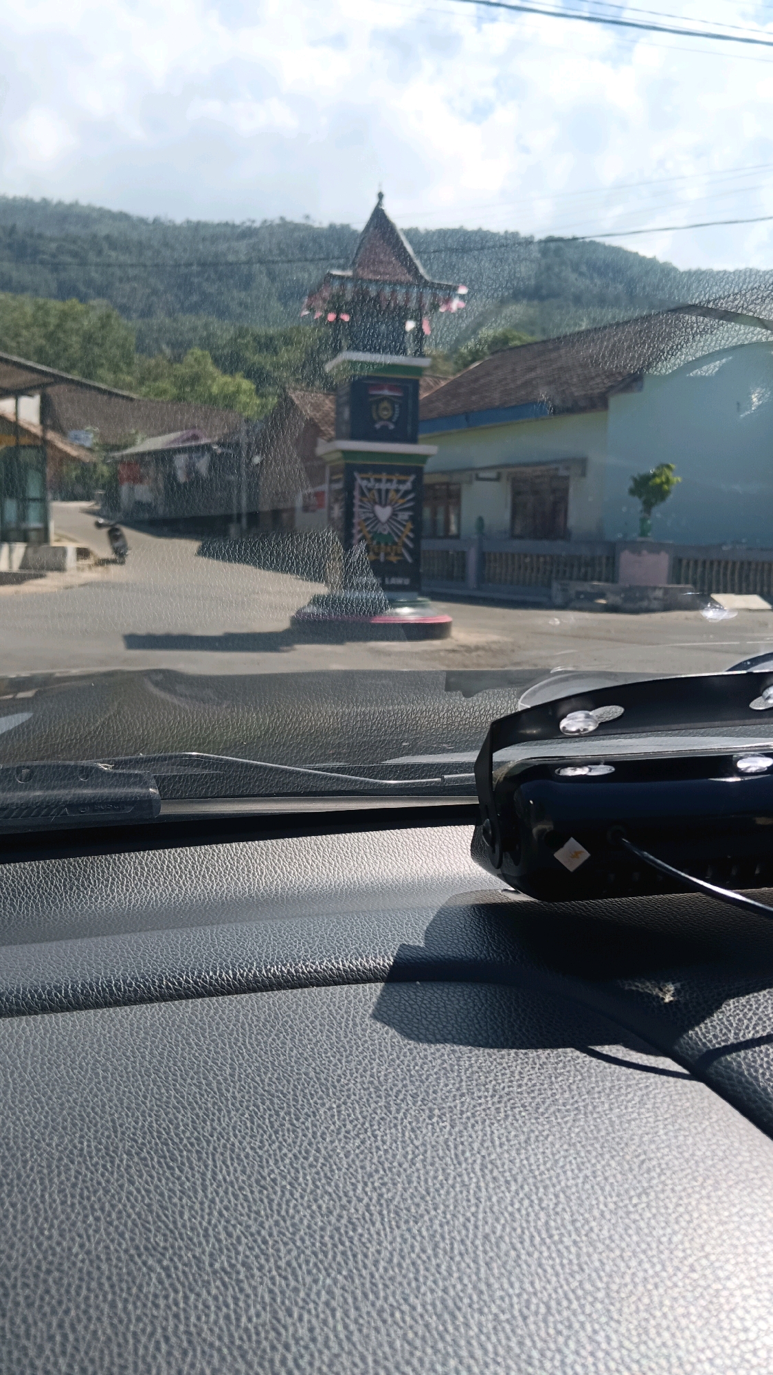 The image, taken from inside a vehicle, shows a view through the windshield of a street in Girimulyo, Indonesia. A prominent landmark, a decorative pillar with a traditional roof structure, stands in the middle of the road. The pillar is adorned with flags and symbols, suggesting a cultural or governmental significance. Surrounding the pillar are buildings with tiled roofs, typical of the region. Lush green hills form the backdrop under a partly cloudy sky. In the foreground, the textured dashboard of the car is visible, along with a black electronic device mounted on the windshield. The scene appears to be a daytime shot, with sunlight casting shadows.