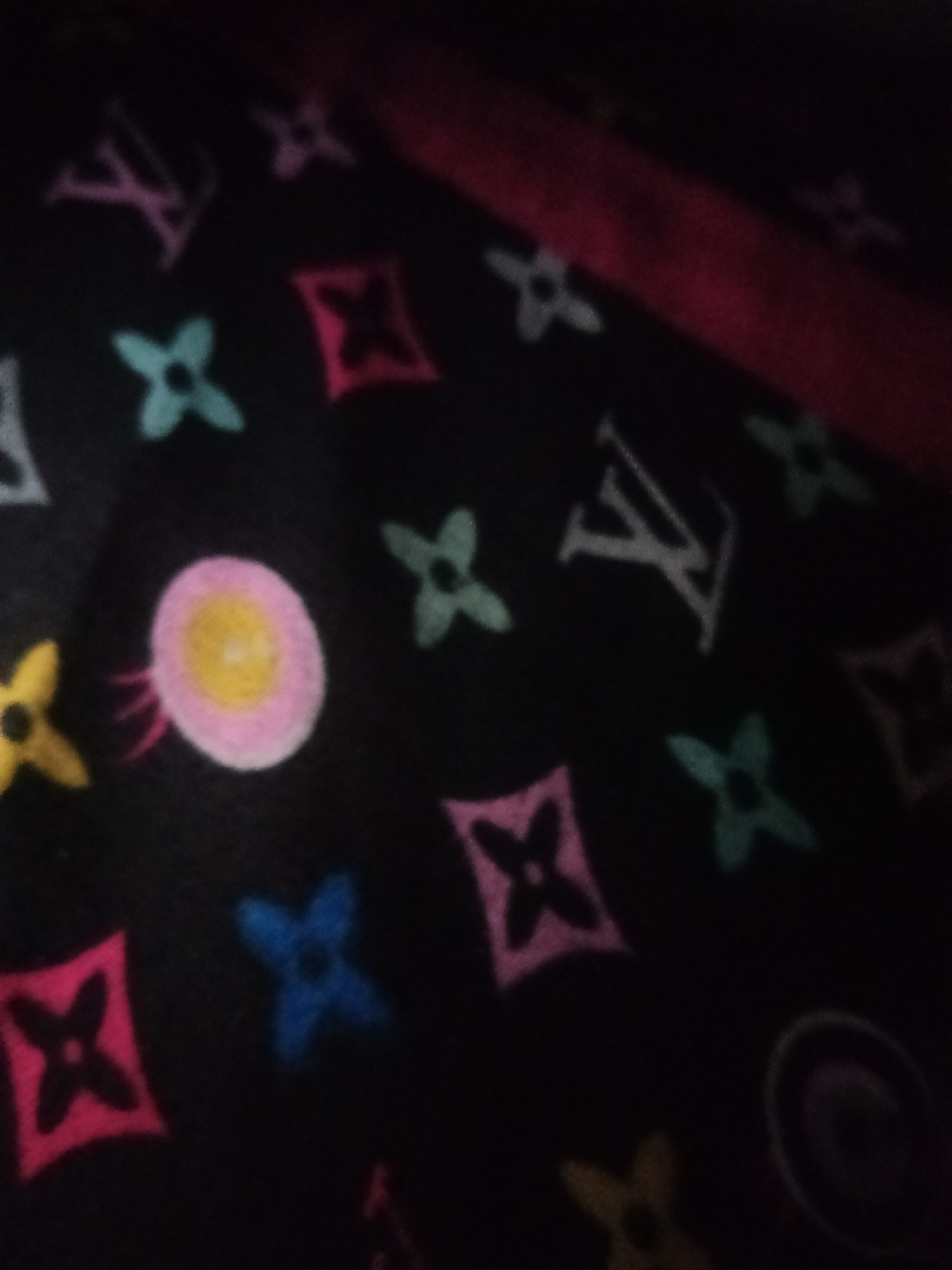 The image is a close-up, blurry photograph of a black fabric with a repeating pattern of colorful shapes and letters. The shapes are abstract and appear to be stylized flowers or four-pointed stars in various colors including pink, teal, yellow, and blue. There are also white letters that resemble "LV," likely a Louis Vuitton monogram pattern. The fabric is folded or draped, with a red, slightly reflective surface visible at the top, suggesting it might be part of a bag or garment. The image appears to have been taken indoors, with low lighting conditions contributing to the blurriness and lack of sharp detail. There is no indication of any specific location in Jalingo, Nigeria.