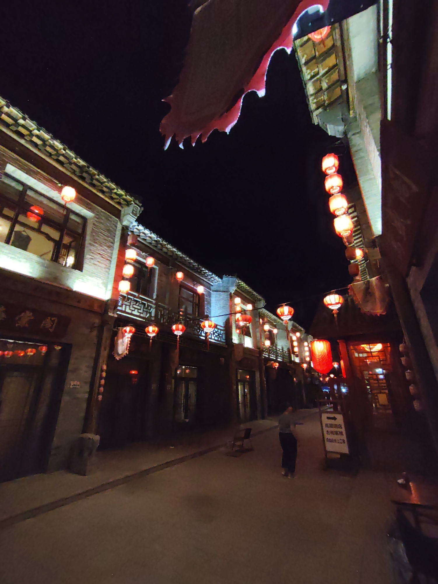 The image depicts a nighttime street scene in Lindian, China, characterized by traditional architecture and illuminated by numerous red lanterns. The street appears to be paved, with buildings lining both sides. The architecture suggests an older style, with multi-story structures featuring visible windows, balconies, and ornate eaves. Red lanterns are strung along the street and hung from the buildings, casting a warm, festive glow. One building on the left has traditional signage with Chinese characters. On the right side of the street, a white sign with Chinese characters and an arrow is visible. A single person, dressed in dark clothing, is walking away from the camera down the street. The overall atmosphere is one of a cultural district or historic area lit up for evening.