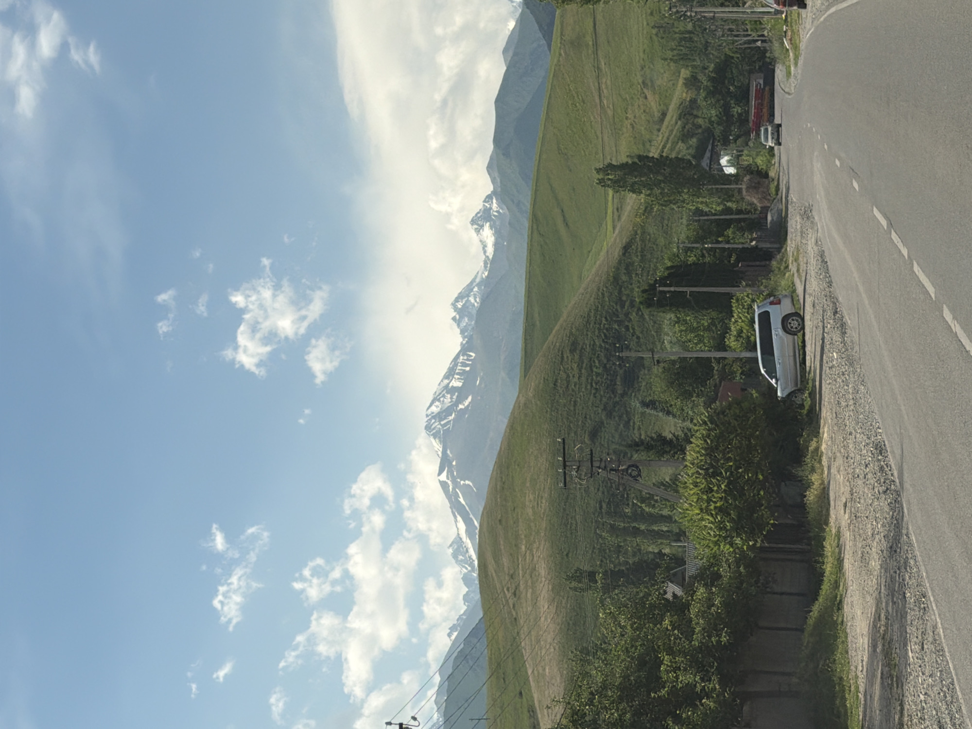 An outdoor, eye-level, full shot captures a paved road leading towards a distant mountain range. The road curves to the right, with a white van parked on the left shoulder. A low concrete fence lines the left side of the road, bordered by green trees. Further back, rolling green hills are interspersed with trees, leading to a majestic mountain range with snow-capped peaks under a blue sky with scattered white clouds. Power lines and poles are visible throughout the scene.