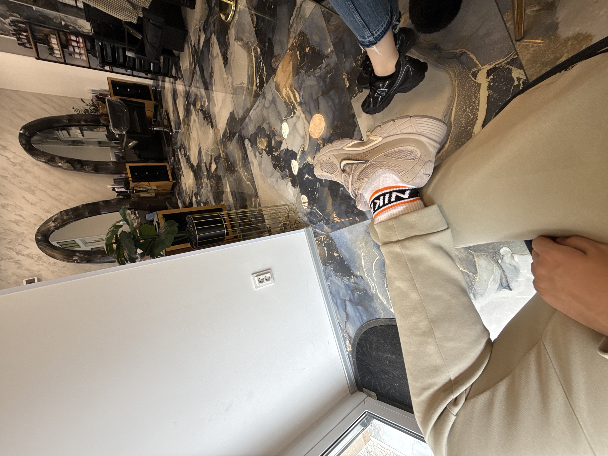 A first-person view captures a person wearing beige pants and a beige sneaker with orange and black striped socks, in what appears to be a commercial interior space. The floor has a marble-like pattern with gold veining. In the background, there is a salon-like setup with mirrors, black furniture, and what appears to be hair products displayed on shelves. Another person can be seen wearing jeans and black sneakers. This photo seems to be taken inside a beauty salon somewhere in Plovdiv, Bulgaria. A white wall with a power outlet is also visible.