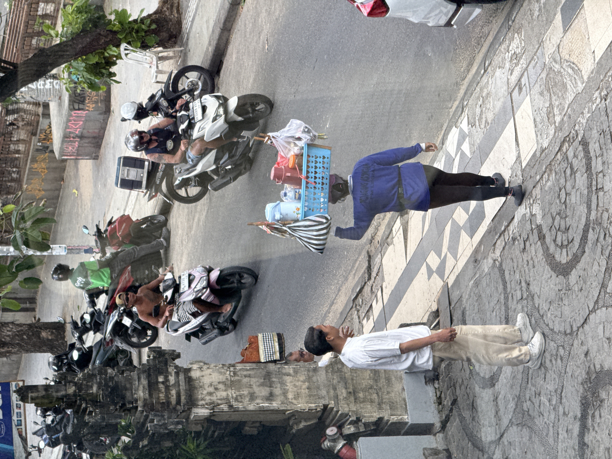The image shows a street in Kuta, Indonesia, with several parked motorcycles along the side of the road. A man in a blue shirt is walking on the sidewalk carrying a basket of goods. A second man in a white shirt stands looking up. The scene includes buildings, street pavement, parked motorcycles, and pedestrians.