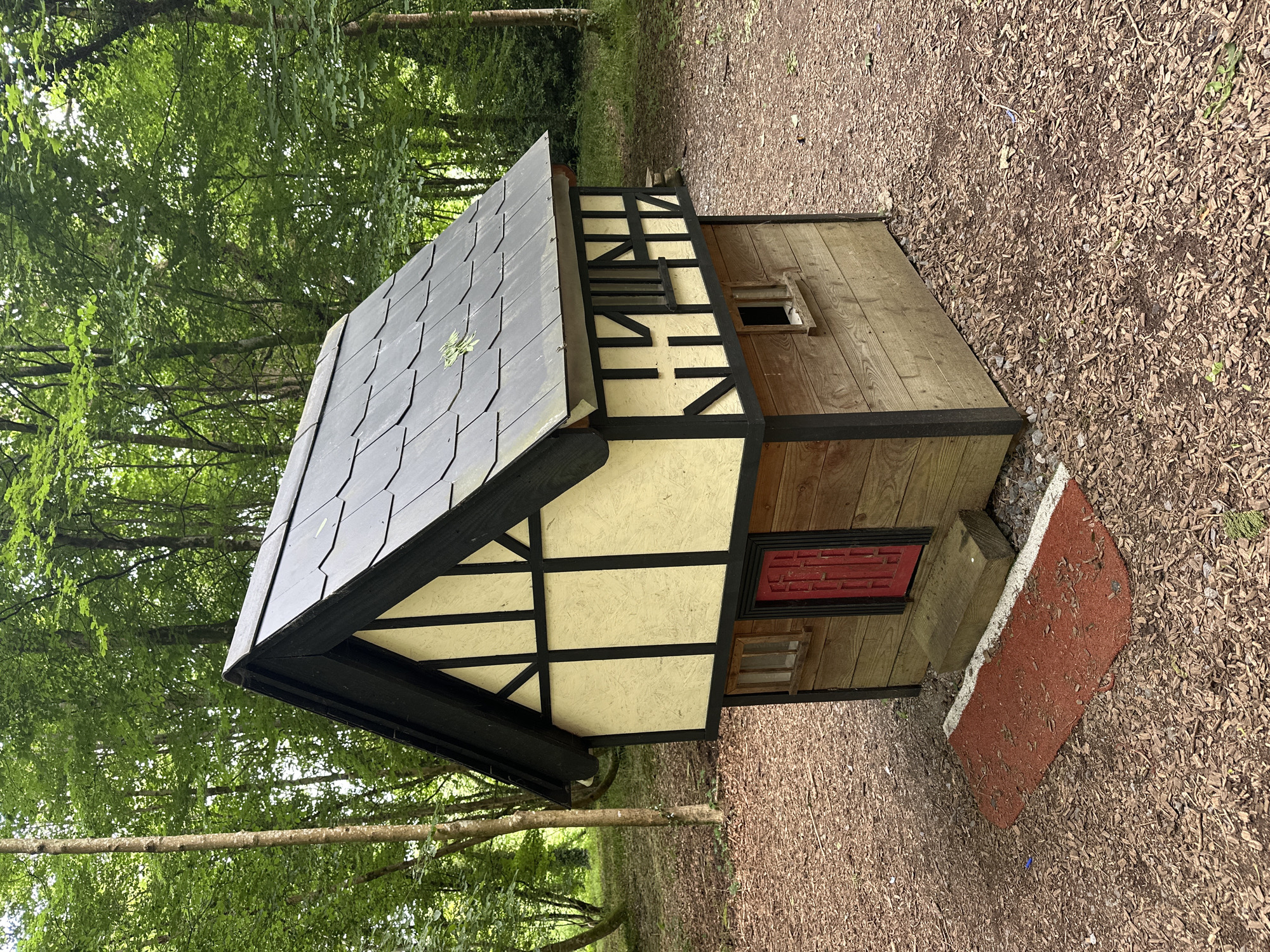 A small, Tudor-style playhouse sits on a woodland floor covered in wood chips and fallen leaves. The playhouse has a dark gray shingled roof, black trim, and cream-colored walls with black timber framing details. It features a red door, two small windows on the front, and a smaller window on the right side. A wooden step leads to the red door, and a red doormat with white edging is placed in front of it. The playhouse is surrounded by trees with green foliage. The scene is likely located in the grounds of Castlecomer Estate in County Kilkenny, Ireland, known for its woodland walks and attractions.