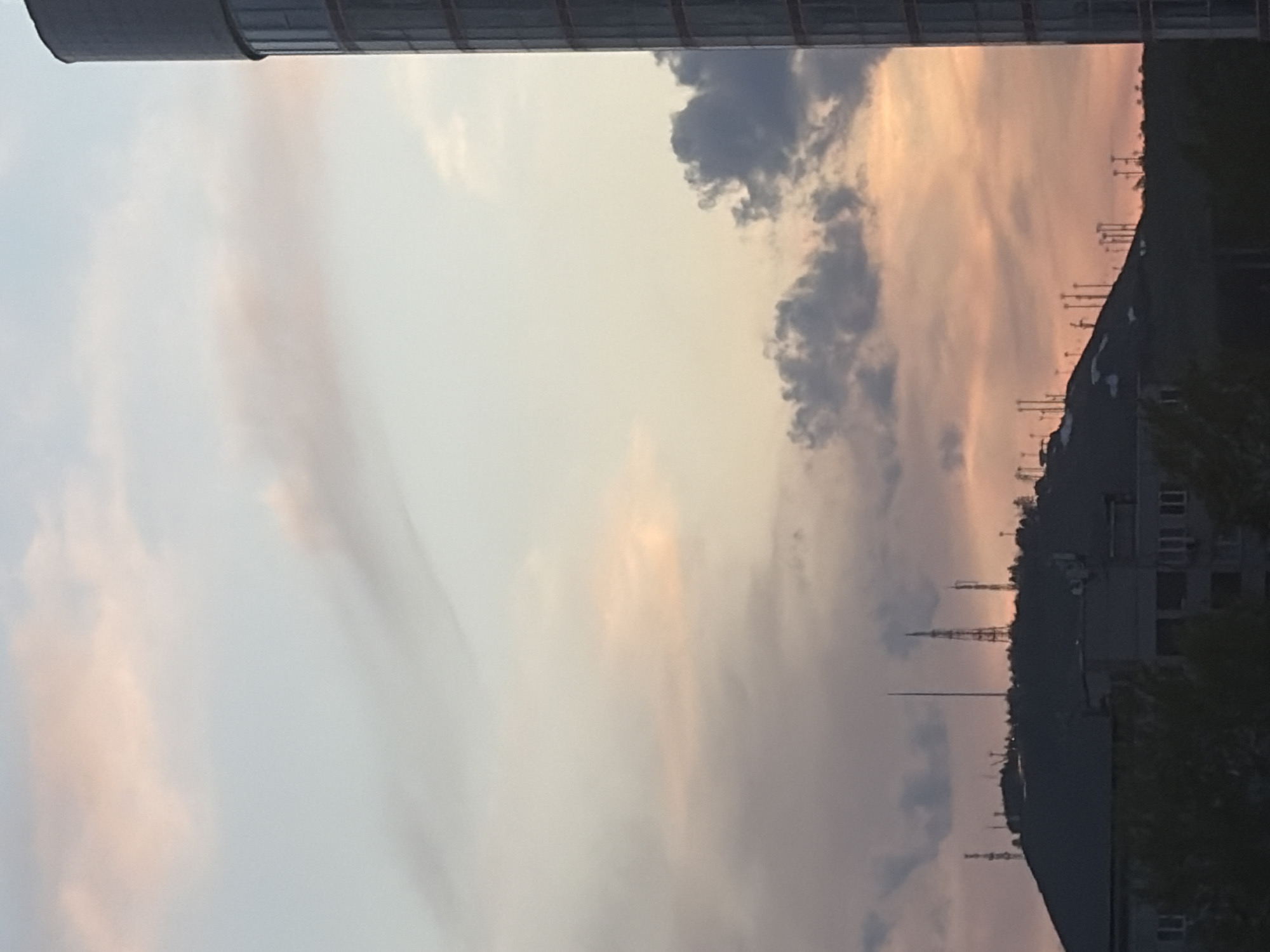 The image depicts a sunset view with city elements. The sky exhibits colors of gray, peach, and blue, with cloud formations. Silhouetted against the sky are buildings and structures, including what appear to be transmission towers. The location is in the city of Krasnoyarsk, Russia. A vertical metallic cylindrical structure is present in the upper left corner.