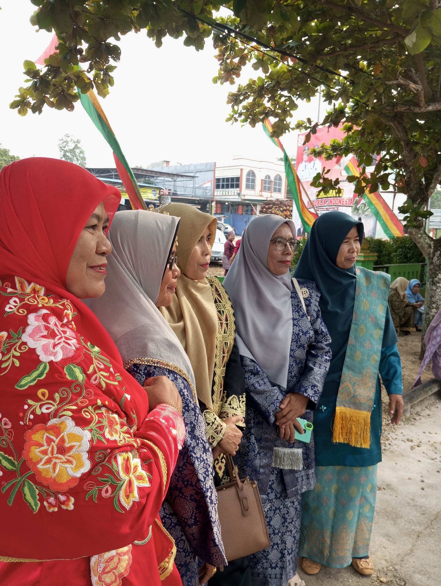 Five women are standing in a line outdoors in what appears to be the city of Pematang Reba, Indonesia. The woman on the far left wears a bright red hijab with intricate floral embroidery. Next to her, a woman in a light gray hijab stands beside another woman in a tan hijab. The fourth woman from the left wears a dark teal hijab and a teal tunic with a patterned scarf draped over her shoulder. The fifth woman, partially visible, wears a dark top with gold embellishments. The women are all facing to the right and appear to be posing for a photo. In the background, there are buildings, trees, and a banner with text in Indonesian. The ground is a mixture of sand and concrete. The overall scene suggests a cultural event or gathering.