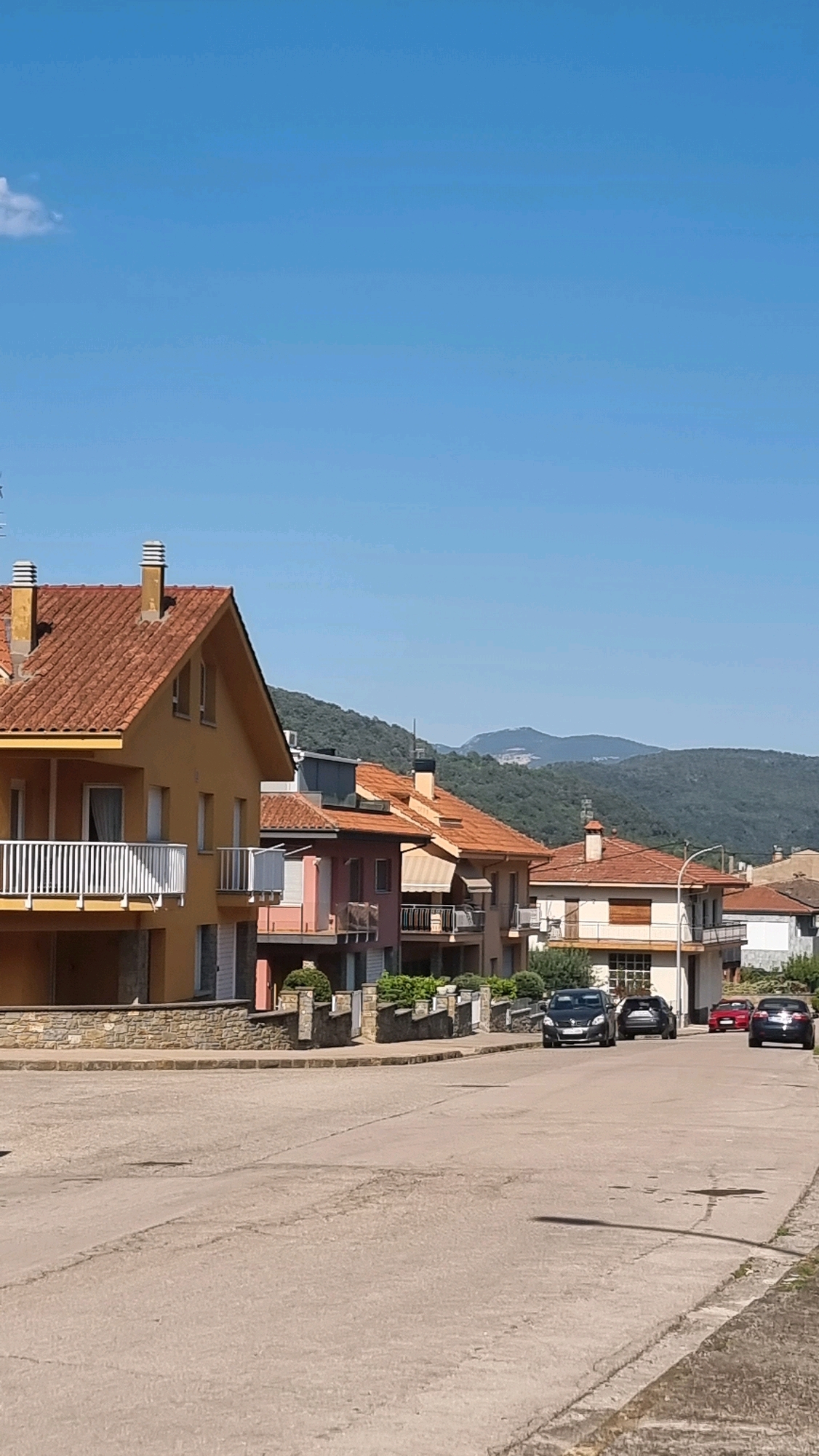 The image depicts a residential street in Sant Joan les Fonts, Spain, on a clear day with a bright blue sky. The street is paved with asphalt and shows some signs of wear. Along the street, there is a row of multi-story houses, characterized by terracotta tile roofs, stucco exteriors in various colors (including yellow, pink, and beige), and balconies with white railings. The houses appear to be of modern construction. Greenery, including hedges and bushes, is visible in front of some of the properties.
In the foreground, the street is empty except for some slight surface irregularities and a long shadow cast by an unseen object, likely a lamp post. Further down the street, several cars are parked or driving. These include a dark grey sedan, a lighter colored sedan behind it, a red car, and a dark blue or black sedan at the far end of the visible street. In the background, a lush, green, tree-covered mountain range rises against the sky, suggesting a natural landscape surrounding the town. A faint silhouette of another mountain range can be seen further in the distance. The overall impression is of a peaceful, suburban or semi-rural environment.