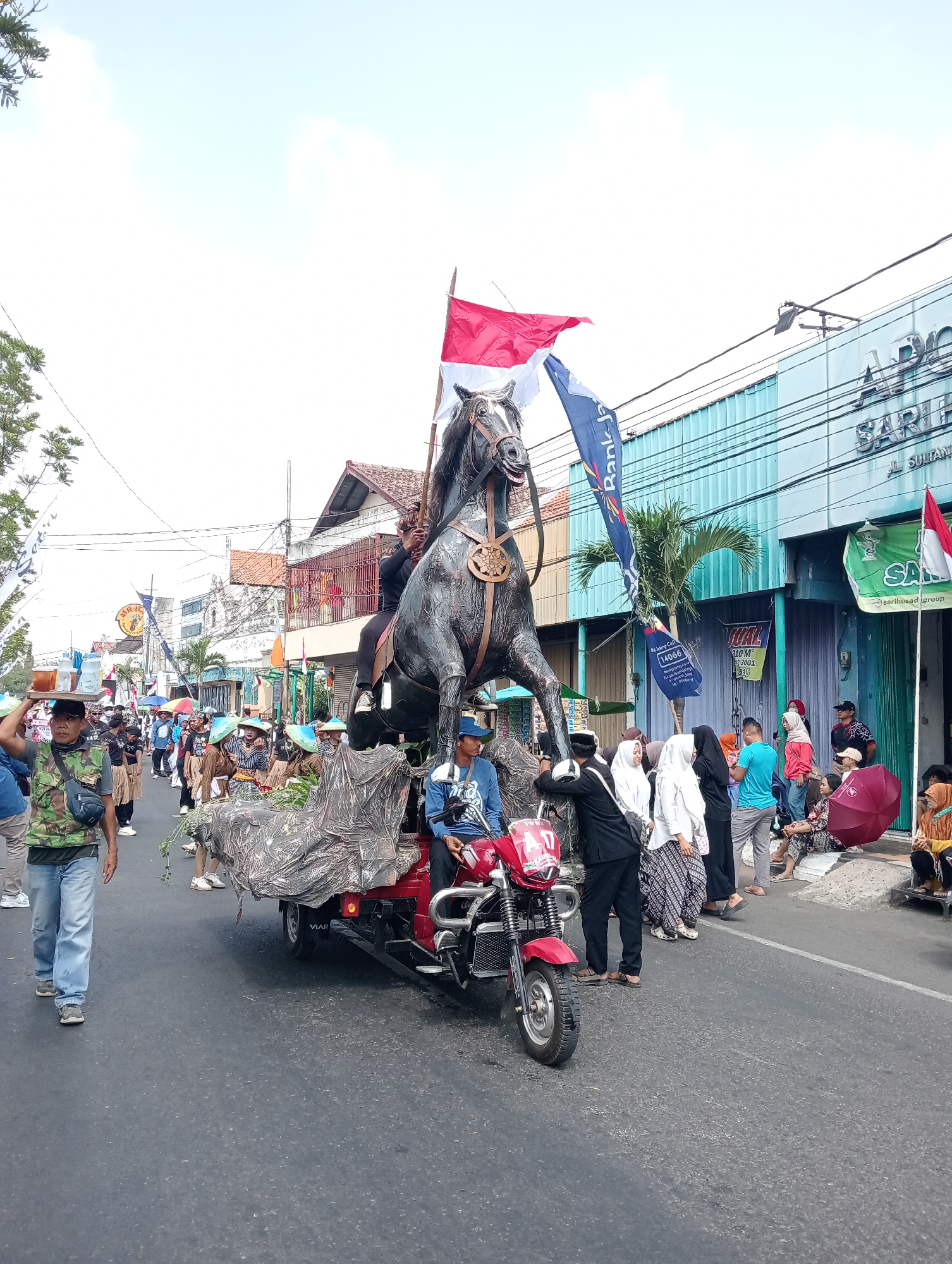 This image depicts a parade or festival in the city of Bintoro, Indonesia. A large, dark statue of a horse is mounted on a red three-wheeled vehicle, which is decorated with camouflage-patterned fabric. Two individuals are positioned on the vehicle, one appearing to control the statue and another seated. A procession of people, some in traditional attire and others in casual clothing, follows behind. Indonesian flags, red and white, are prominently displayed. The scene takes place on a paved street lined with buildings, some of which appear to be businesses with signage. The sky is overcast.