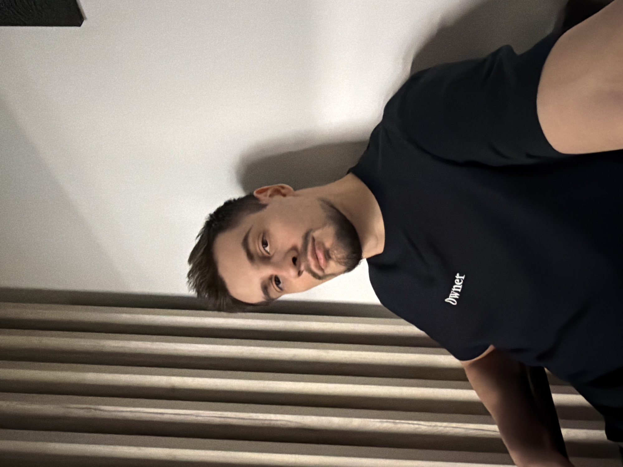 A man stands indoors in front of horizontal blinds. He wears a dark t-shirt with the word "owner" printed on the left sleeve. A shadow is cast on the white wall behind him. The location cannot be confirmed as Krasnoyarsk, Russia from this image.