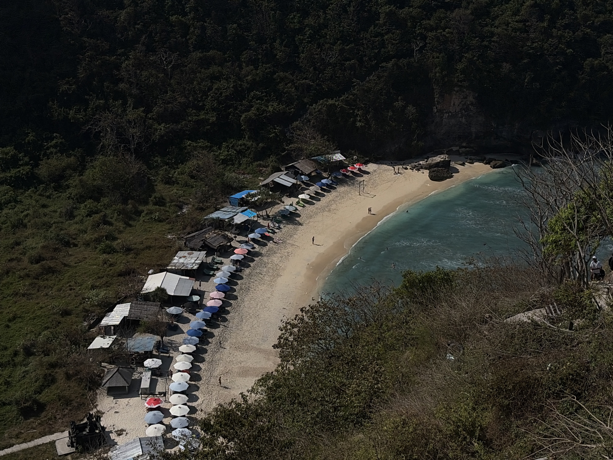 This is an aerial, high-angle view of a resort beach area on Nusa Penida, Indonesia. The beach is a crescent of light-colored sand meeting turquoise water. Along the edge of the sand, adjacent to a lush, dark green, forested hillside, are numerous small structures that appear to be beach huts or bungalows. In front of these structures, arranged in neat rows, are many colorful beach umbrellas. Several people are visible on the beach, some walking and some near the water. The overall scene depicts a tranquil, tropical beach environment.