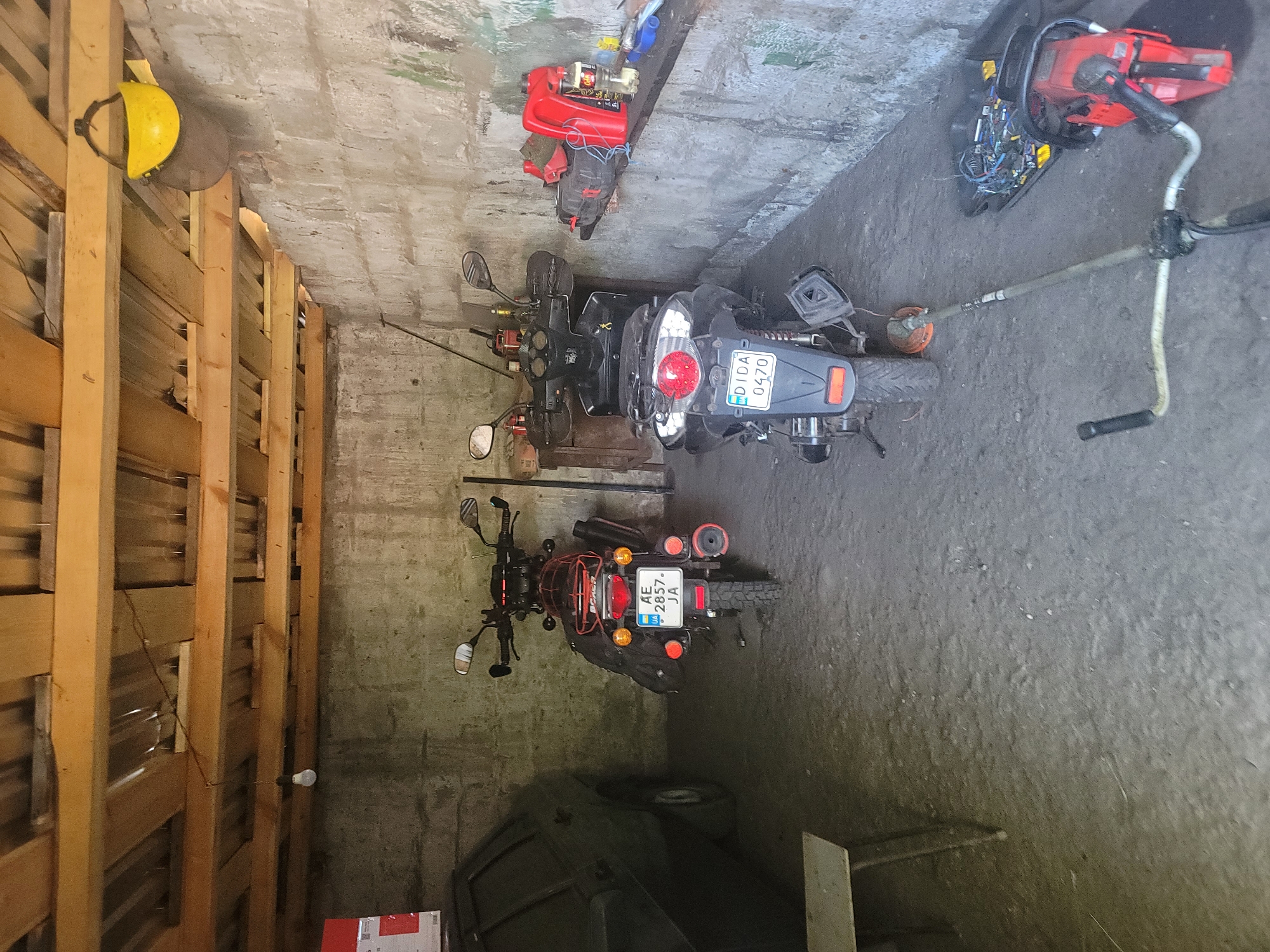 In a garage in Vovkivka, Ukraine, two motorcycles, a car, a brush cutter, and a chainsaw are visible. The garage has a concrete floor and block walls, with a wooden roof structure. One motorcycle is a black scooter with the license plate "DIDA 0470". The other is a red dirt bike with the license plate "AE 2857 JA". A dark-colored car is parked to the left of the dirt bike. A brush cutter lies on the floor in front of the scooter, and a red chainsaw is also on the floor to the right. A workbench with various items, including red gas cans and tools, is against the back wall. A yellow hard hat is hanging from a ceiling beam.