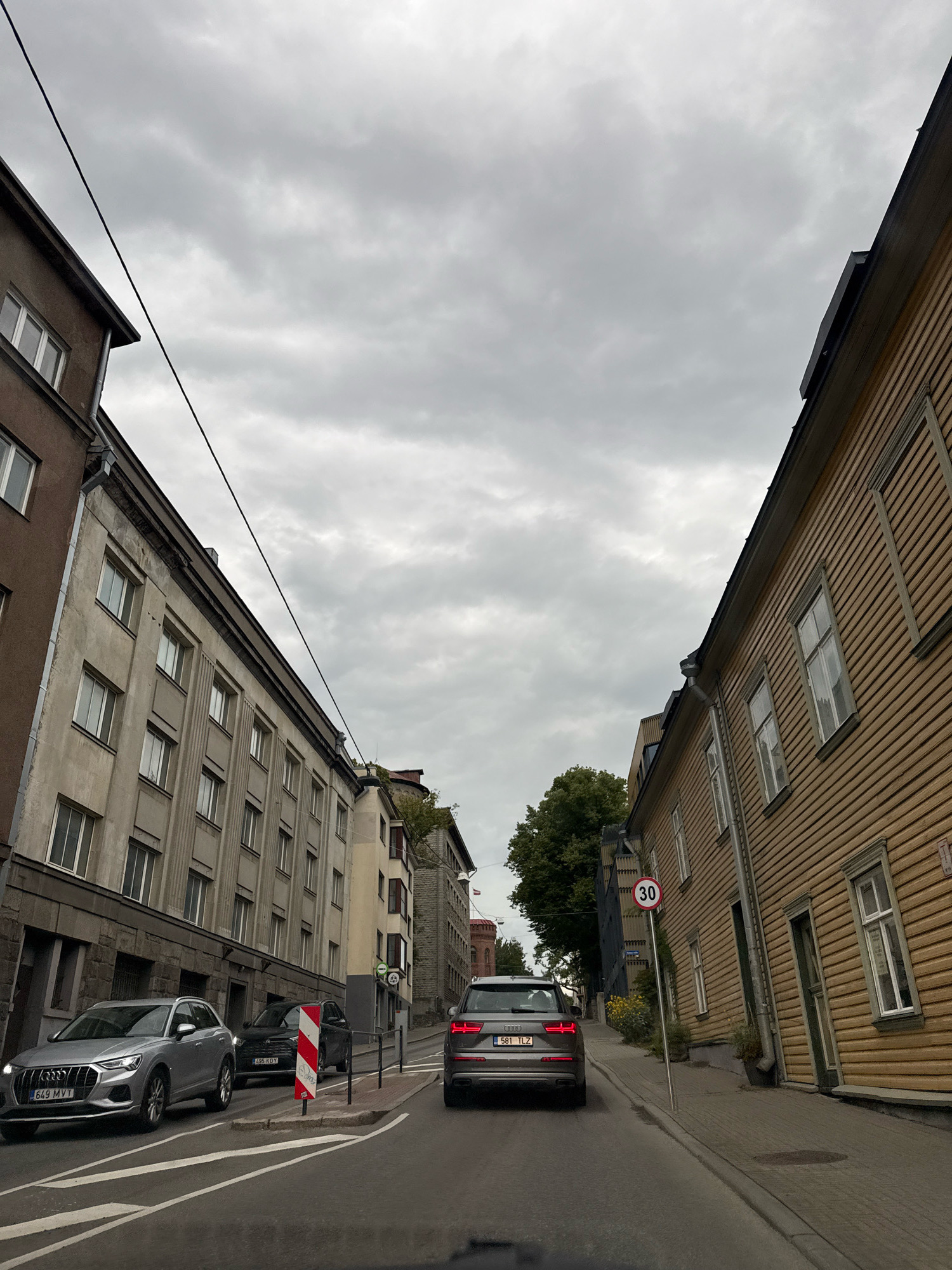The image depicts a street scene in Tallinn, Estonia. A grey Audi SUV is in the foreground, facing away from the camera. To the left, a silver Audi Q3 is parked. The street is lined with buildings on both sides, with a mix of architectural styles. On the right, a yellow wooden building with white-framed windows stands prominently. On the left, a taller, grey apartment building with multiple windows is visible. The sky is overcast and grey. A speed limit sign indicating "30" is present on the right side of the street. The scene appears to be a residential or mixed-use urban area.