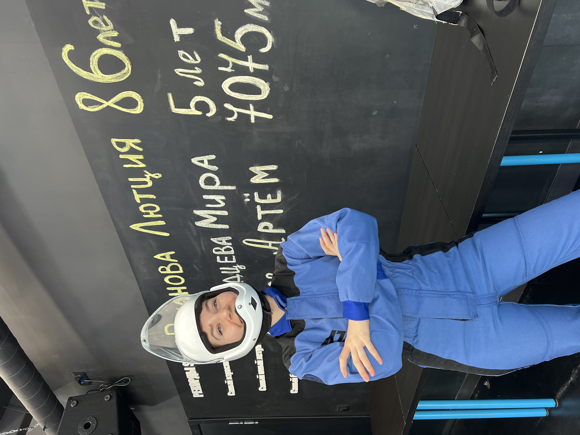 A woman wearing a blue jumpsuit and a white helmet stands in front of a black wall. The wall has white and yellow writing on it, with the words "RECORDS" and "The oldest" visible. The writing also mentions the names "Lyutzia", "Mira", and "Artem", along with ages and a number, potentially indicating a height. The photograph was taken at AEROSIM Izhevsk, located in the city of Izhevsk, Russia.