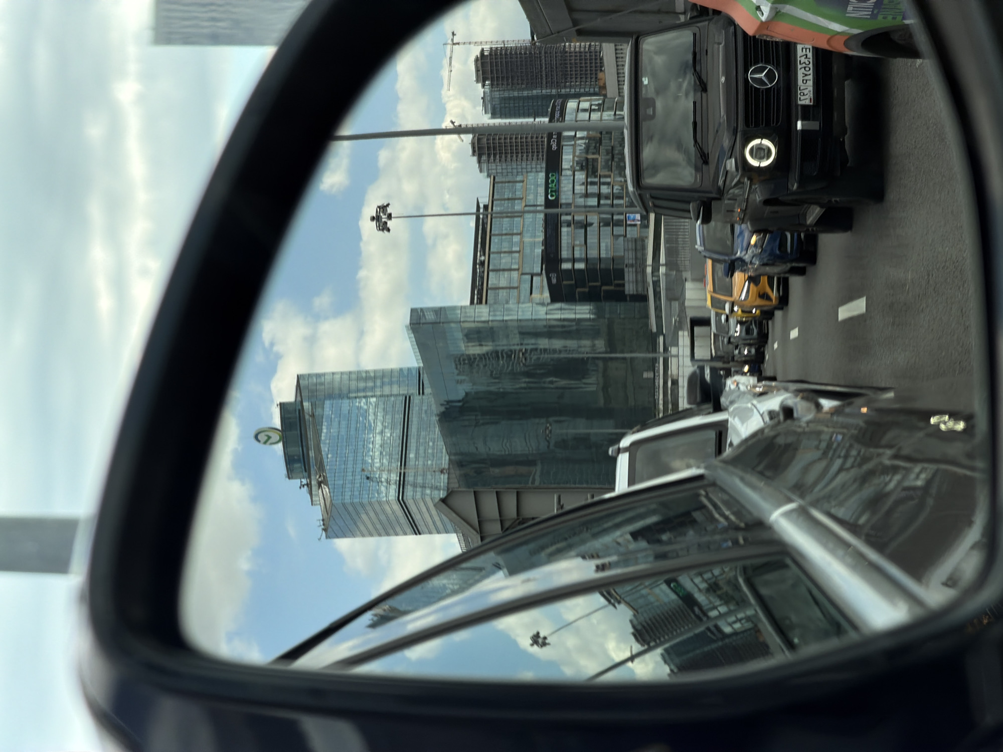 The image captures a view in Moscow, Russia, as reflected in a car's side mirror. The mirror shows a cityscape featuring modern buildings with reflective glass facades, including a building with a "VTB Arena park" logo on top. Construction cranes are visible in the background, suggesting ongoing development. The reflection also shows a line of vehicles on the road adjacent to the car, including a black Mercedes G-Class with its license plate partially visible, and cars of different colors, like yellow, blue, and silver. The sky is partly cloudy. The black car mirror dominates the frame.
