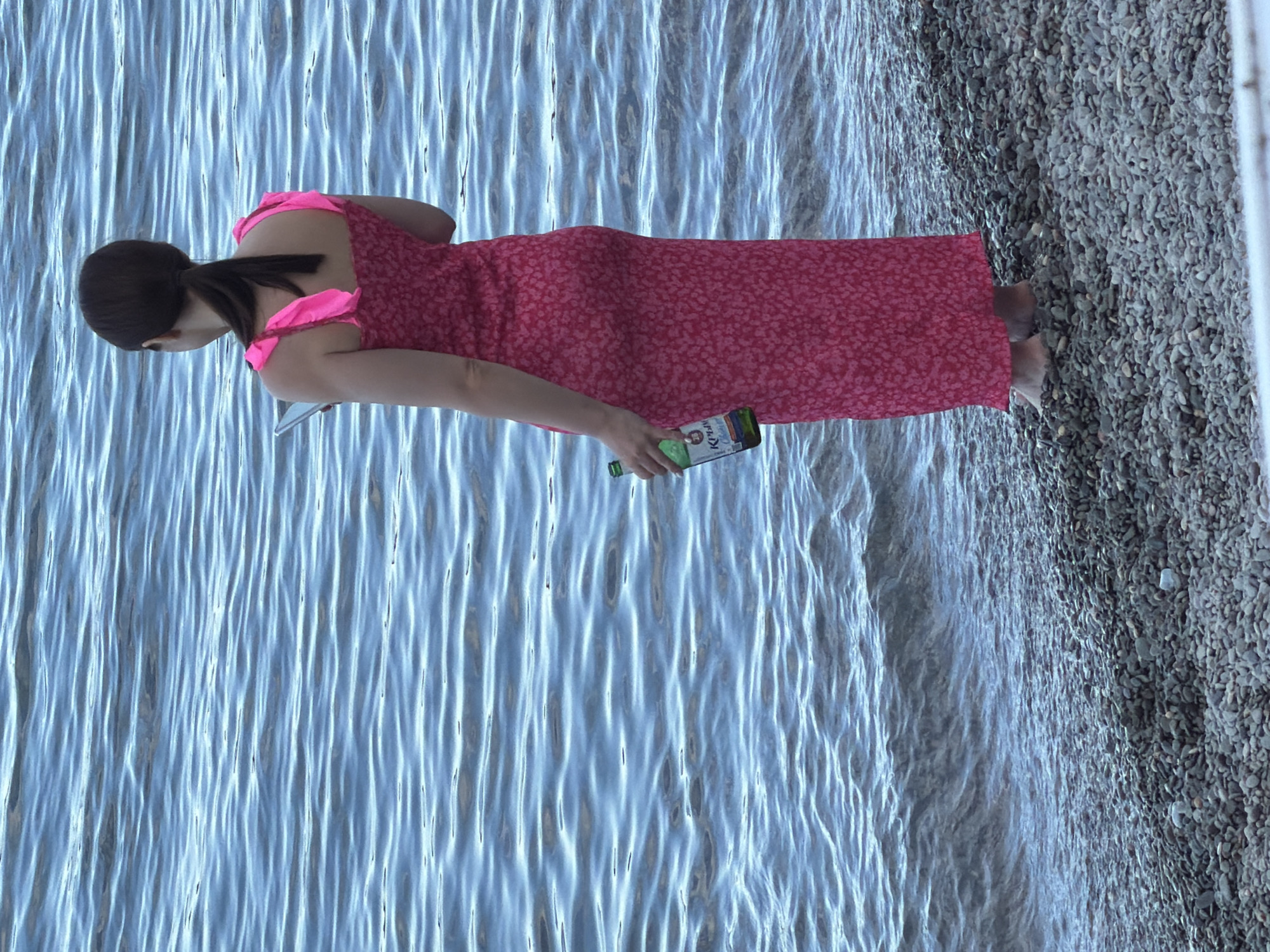 A woman with dark hair pulled into a ponytail stands on a pebbled shore, facing away from the camera. She is wearing a long, bright pink, floral-patterned dress with ruffled straps. In her right hand, she holds a green glass bottle with a partially visible label. Her left hand is extended slightly forward, holding a flat, silver object, possibly a phone or tablet. The water behind her is a deep blue, with gentle ripples. The scene appears to be a natural setting along the coast of Partenit, Ukraine.
