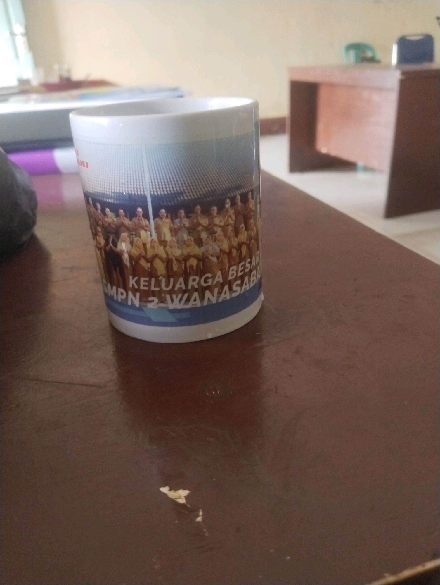 The image shows a white ceramic mug with a picture of a group of people on it, along with text that reads "KELUARGA BESAR" and "SMPN 2 WANASABA". The mug is placed on a dark brown, polished surface, possibly a table. In the background, there is a desk and chairs, suggesting an indoor environment such as an office or classroom. The lighting appears to be artificial, with the overall scene having a warm tone. The background is slightly out of focus. There is a blurry dark object to the left of the mug, partially obscuring it. The image is cropped to focus on the mug and its immediate surroundings, with the broader environment, including any indication of the city of Kareng Baru, Indonesia, not clearly discernible.