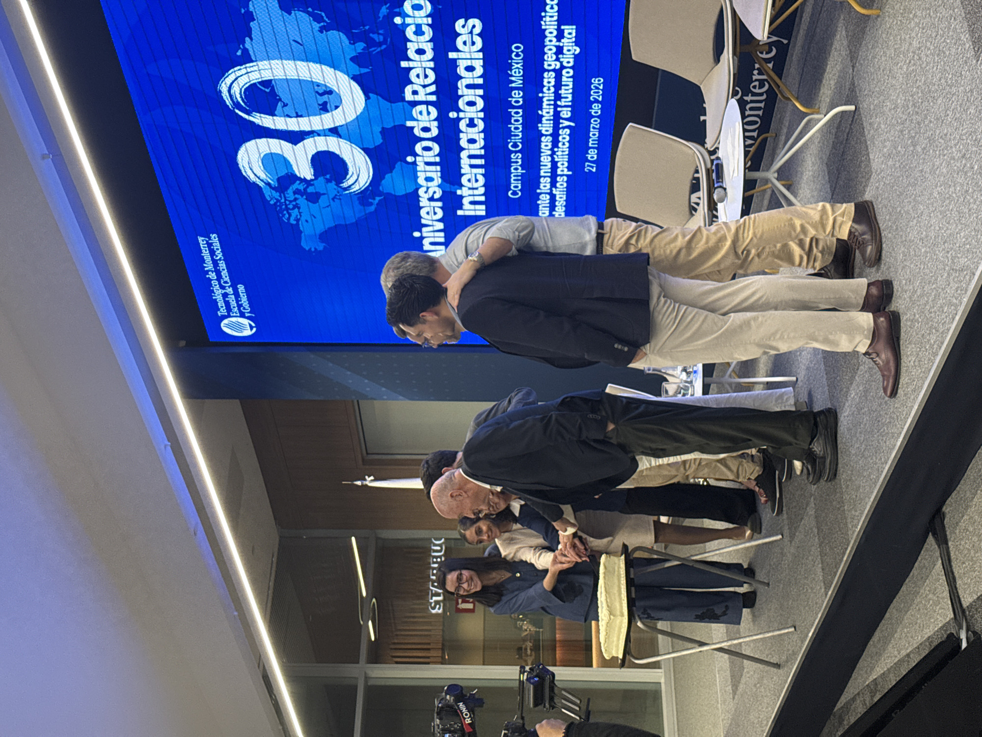 An indoor event is captured at Tecnológico de Monterrey, Campus Ciudad de México, Mexico City, on March 27, 2026, celebrating the 30th Anniversary of International Relations for the School of Social Sciences and Government. Five individuals are centrally positioned on a carpeted stage, with their hands stacked together on a rectangular white cake resting on a collapsible silver stand, performing a symbolic cake-cutting.
To the left of the cake, a smiling woman with glasses in a blue pantsuit participates. An older, bald man in a dark suit stands prominently among the group, also with his hand on the cake. A large blue LED screen in the background displays the event details, including "Tecnológico de Monterrey Escuela de Ciencias Sociales y Gobierno," "30 Aniversario de Relaciones Internacionales," "Campus Ciudad de México," and "27 de marzo de 2026," superimposed over a stylized world map.
On the far left, a video camera on a "RONIN" gimbal is recording the scene. To the right, two men stand on the stage with their backs to the camera, one with an arm around the other's shoulder. A white round side table holding a microphone and papers, along with several beige modern chairs, are situated on the right side of the stage. Through a glass partition in the background, a "STARBUCKS" sign is visible. The environment is well-lit by overhead strip lighting.