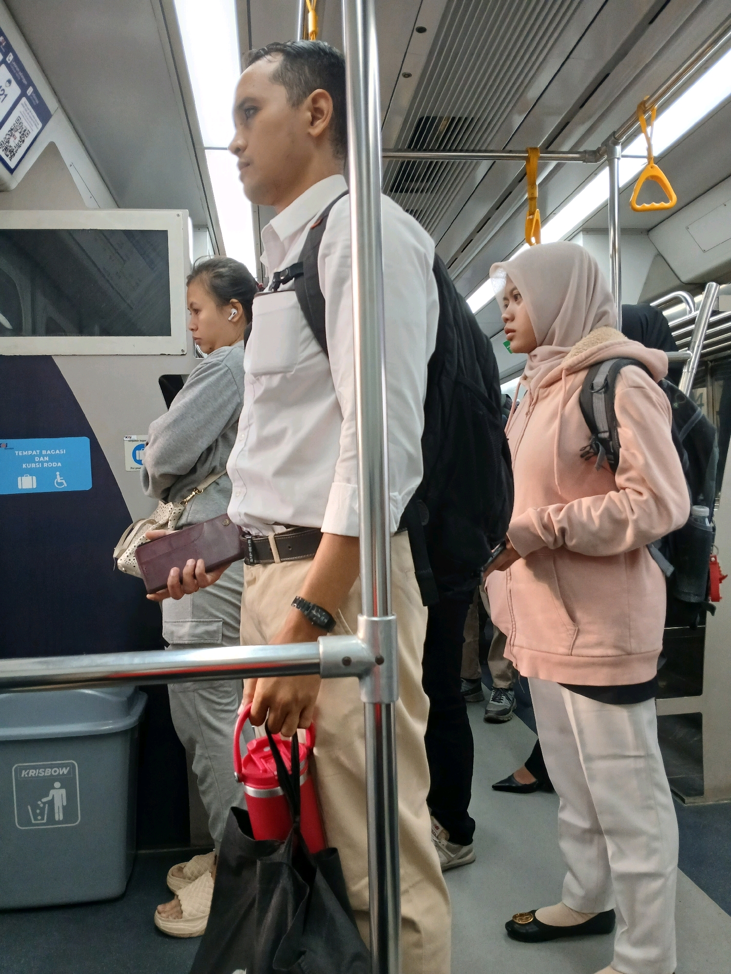 In the City of Medan, Indonesia, an interior view captures several individuals within a train car, illuminated by overhead lights. A man, prominently featured in the foreground, stands facing right, wearing a white collared shirt, tan pants, a black backpack, and a dark watch on his left wrist. He holds a brown wallet and a red thermos with a black bag. To his left, a woman with dark hair, wearing a grey long-sleeved shirt and white earbuds, looks ahead. Further left, a woman in a light pink hijab and a pink hooded jacket with a backpack stands facing right. Other individuals are partially visible in the background. The environment includes metallic poles, yellow and orange handrails, a blue sign with white text and icons indicating luggage and wheelchair space, and a grey trash bin labeled "KRISBOW" with a recycling symbol. The floor is grey.