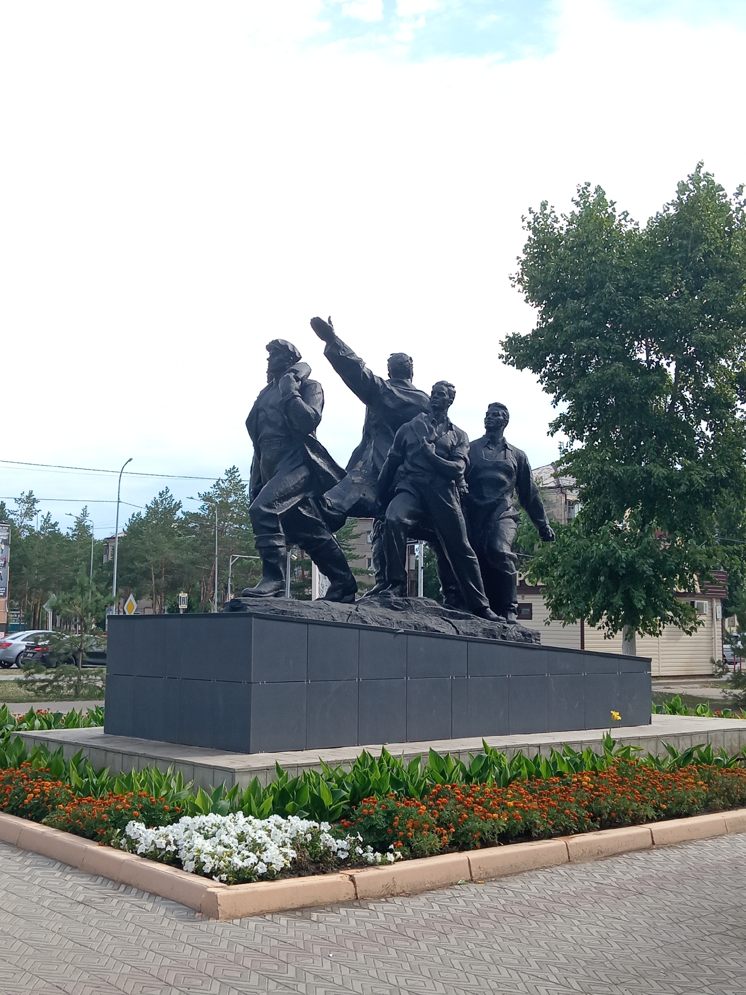 The photograph depicts a monument in the city of Rudny, Kazakhstan. The monument features a dark bronze statue of four men standing on a dark grey rectangular base. The men are depicted in various poses of struggle and leadership, suggesting a historical or revolutionary theme. The front left figure is the tallest, with one arm raised upwards, and the other figures are positioned behind and around him. The monument is surrounded by a landscaped area with green foliage and rows of small orange and white flowers. The ground in front of the landscaped area is paved with light grey interlocking bricks. In the background, there are trees, buildings, streetlights, and a few cars on a road, indicating an urban setting. The sky is overcast with some patches of blue and clouds. The lighting suggests it is daytime.