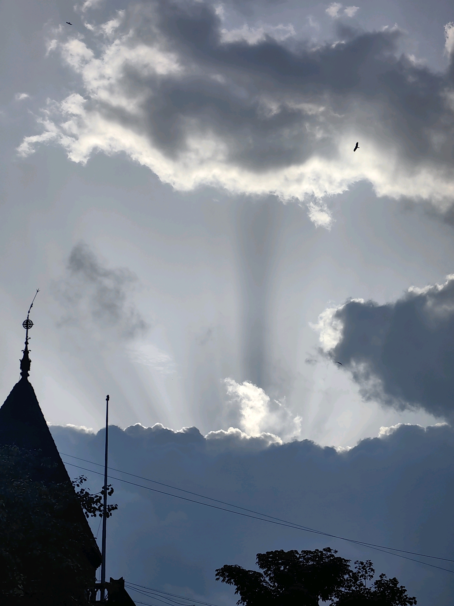 The image depicts a dramatic sky with crepuscular rays filtering through dark, heavy clouds. In the foreground, the silhouette of a building, possibly a church or a historic structure with a spired roof and a decorative finial, is visible on the left. A tall, thin pole, likely a flagpole, stands near the building. The sky is filled with various cloud formations, some dark and ominous, others lighter and catching the sun's rays. Two birds are visible flying in the sky. The overall impression is one of natural beauty and a potentially approaching storm. The image does not contain any identifiable Mumbai landmarks or elements that would definitively place it in Mumbai, India.