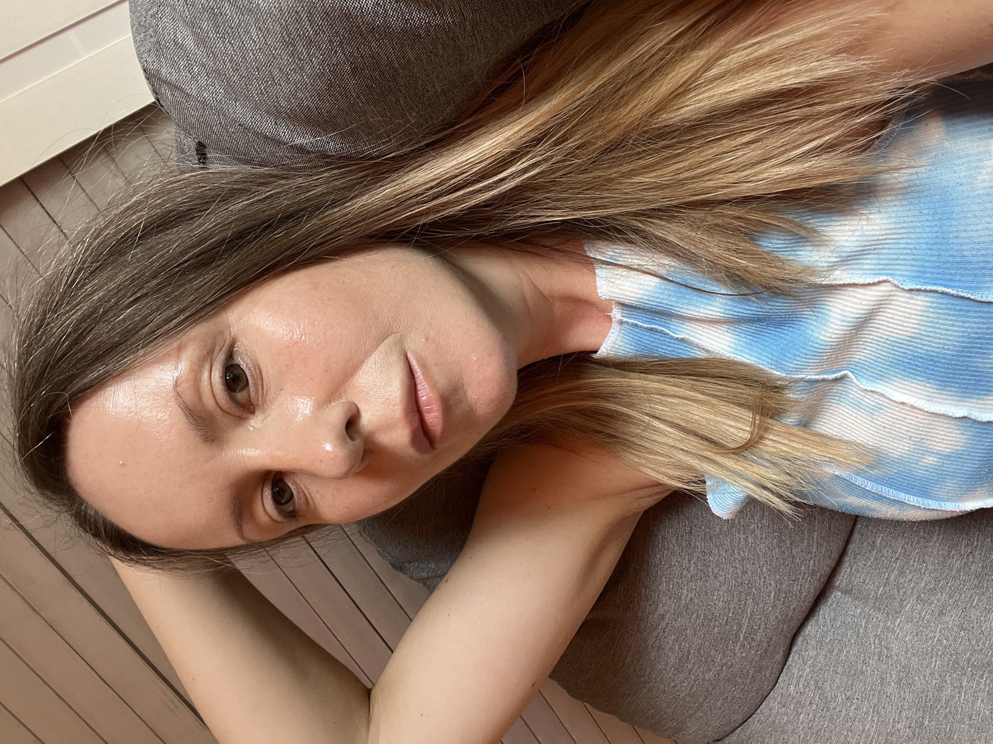 The image shows a close-up, selfie-style shot of a woman with blonde hair, wearing a blue and white tie-dye tank top. She is lying down, with her left arm raised and resting behind her head, and her body is supported by a grey couch. The background appears to be a wooden wall with horizontal planks. There are no discernible landmarks or specific architectural features that would indicate a location in Straubing, Germany. The image focuses on the woman's face, her arm, and the upper portion of her torso. The lighting suggests an indoor setting.