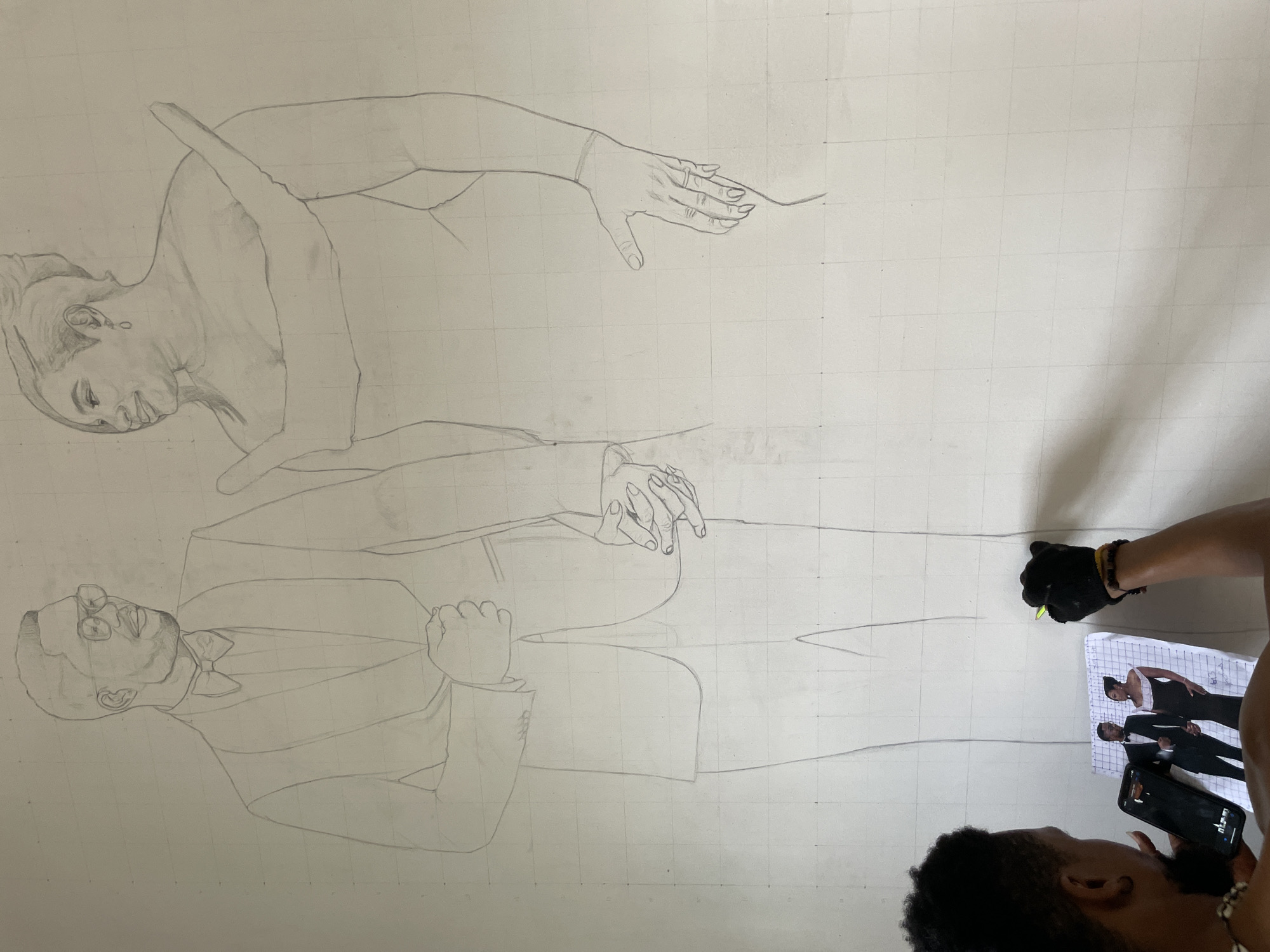 An artist sketches a portrait of a formally dressed couple onto a grid-lined wall. The female figure wears a gown and the male figure wears a suit with a bow tie and glasses. The artist holds a reference photo and uses a pencil to transfer the image. The location appears to be indoors.