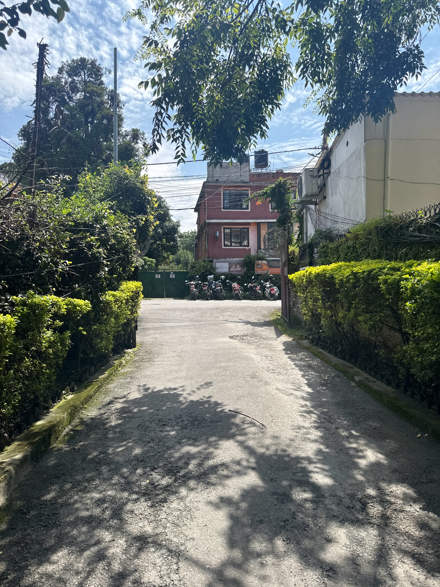 The image captures a sunlit outdoor scene in Lalitpur, Nepal, featuring a pathway leading towards a building complex. The pathway is flanked by well-maintained green hedges on either side. In the background, a red multi-story building is visible, with a white building to its right. Parked motorcycles are lined up in front of a green gate, suggesting a residential or commercial area. The sky is partly cloudy, and dappled shadows from tree leaves are cast across the pathway, indicating a sunny day. The overall impression is one of a peaceful urban environment with abundant greenery.