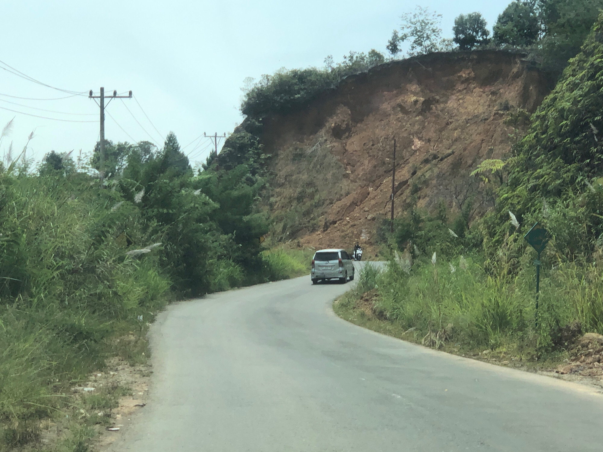 The image shows a winding asphalt road passing through a rural landscape with lush green vegetation and a steep, eroded hillside. A silver minivan is traveling on the road, followed by a motorcycle. Utility poles with wires are visible in the background on the left side of the road. The sky is overcast. The scene appears to be in Bintangpermata, Indonesia, based on the provided context.